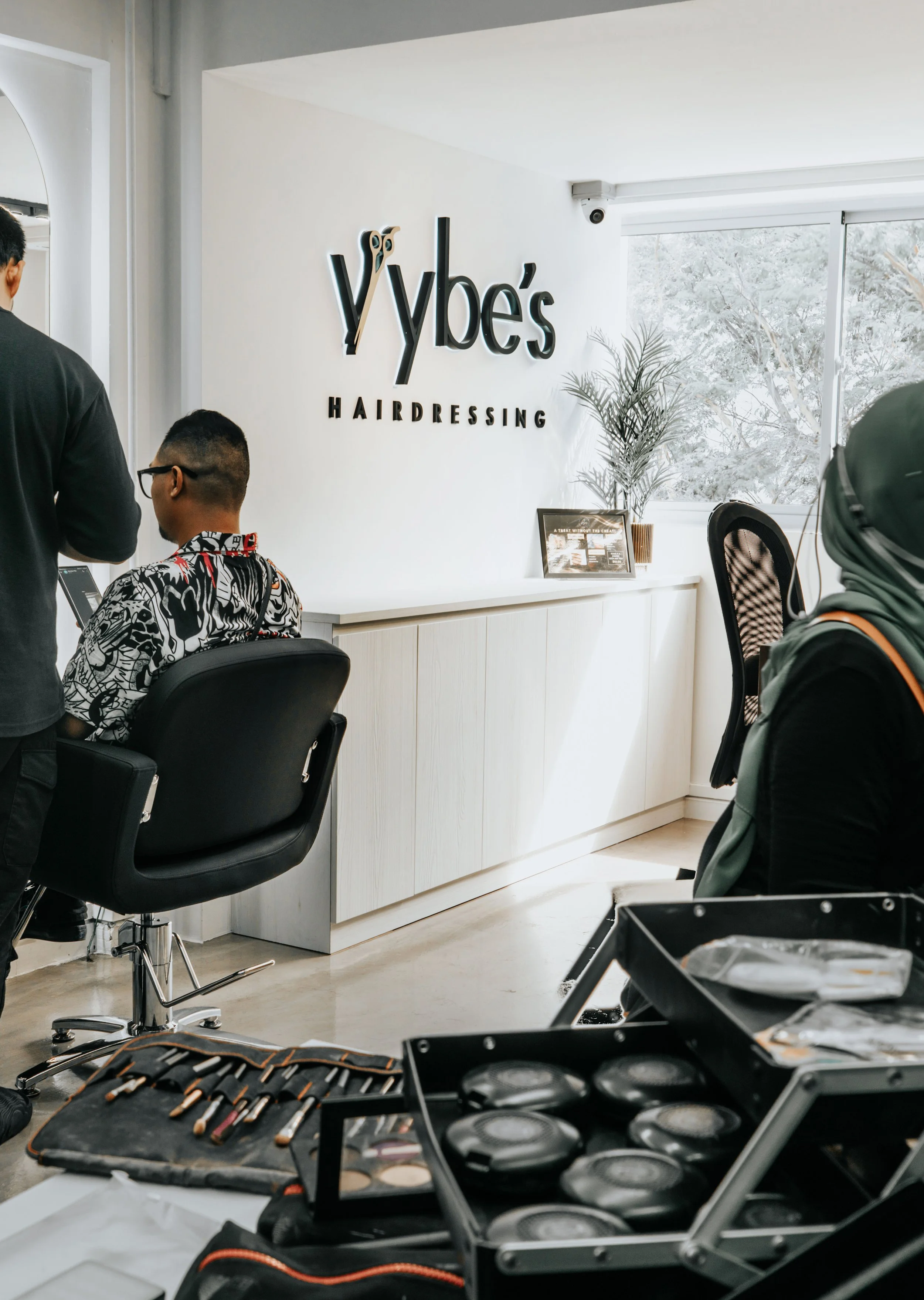 Interior of a salon called Vybe's Hairdressing with some people getting hair services, visible makeup tools, and a large window with trees outside.