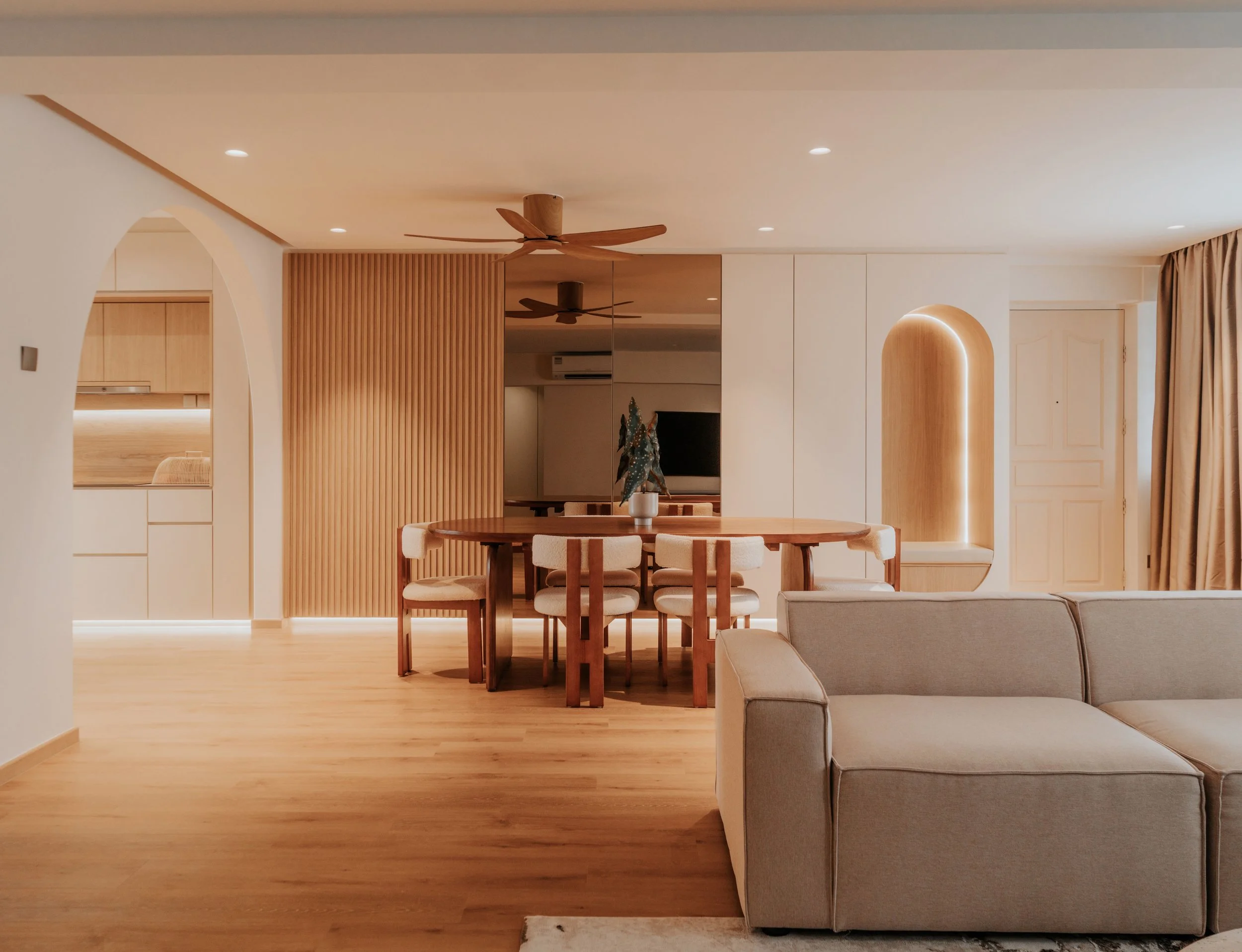 A living room with a beige sofa, a wooden dining table with six chairs, a wall with vertical wooden slats, a mirror, ceiling fans, and a TV in the background. Light-colored hardwood floors and warm ambient lighting are also visible.