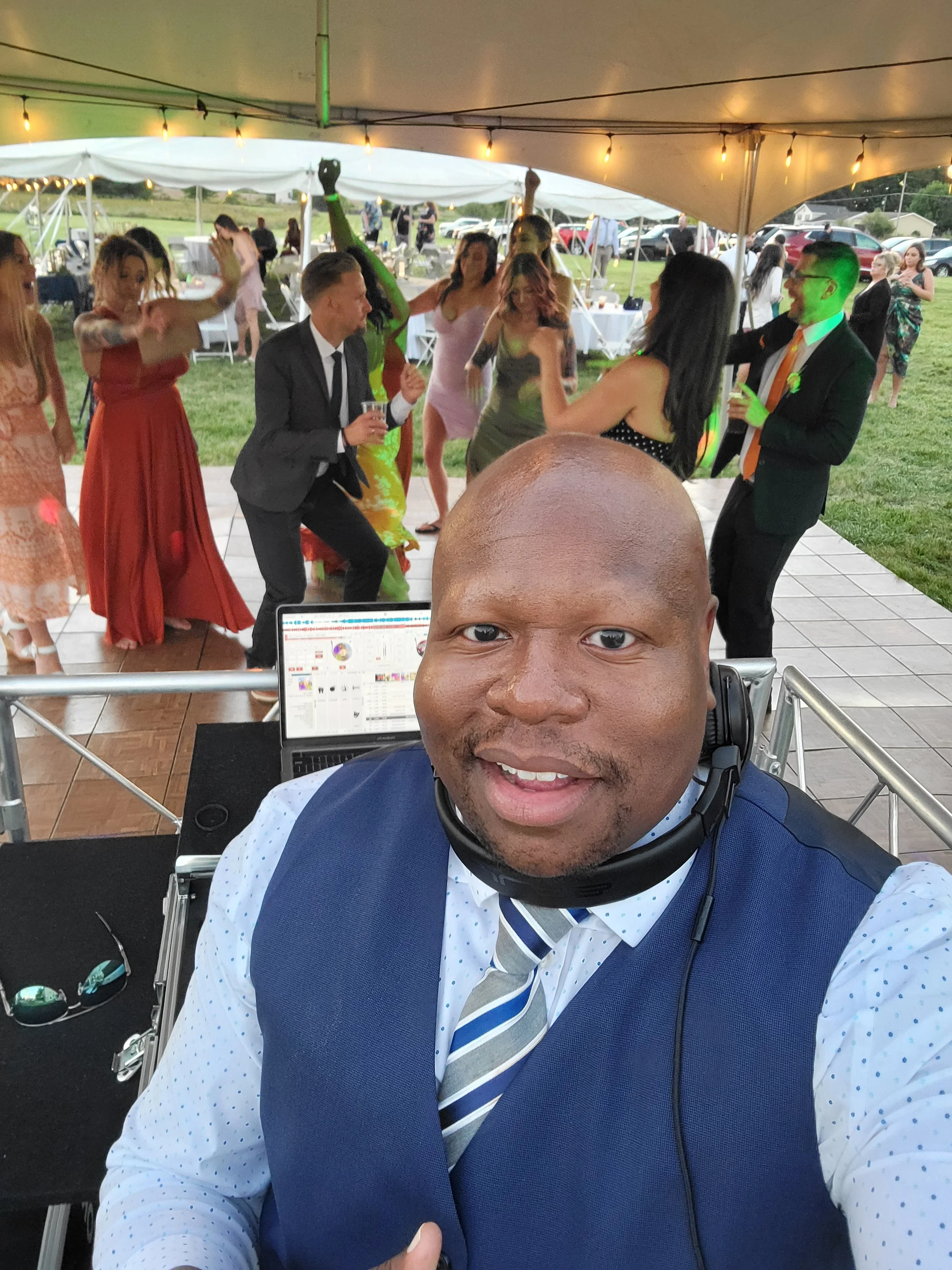 A man wearing a blue vest, dress shirt, and striped tie taking a selfie at a celebration or party under a tent with dancing guests in the background, including people in formal and colorful dresses.