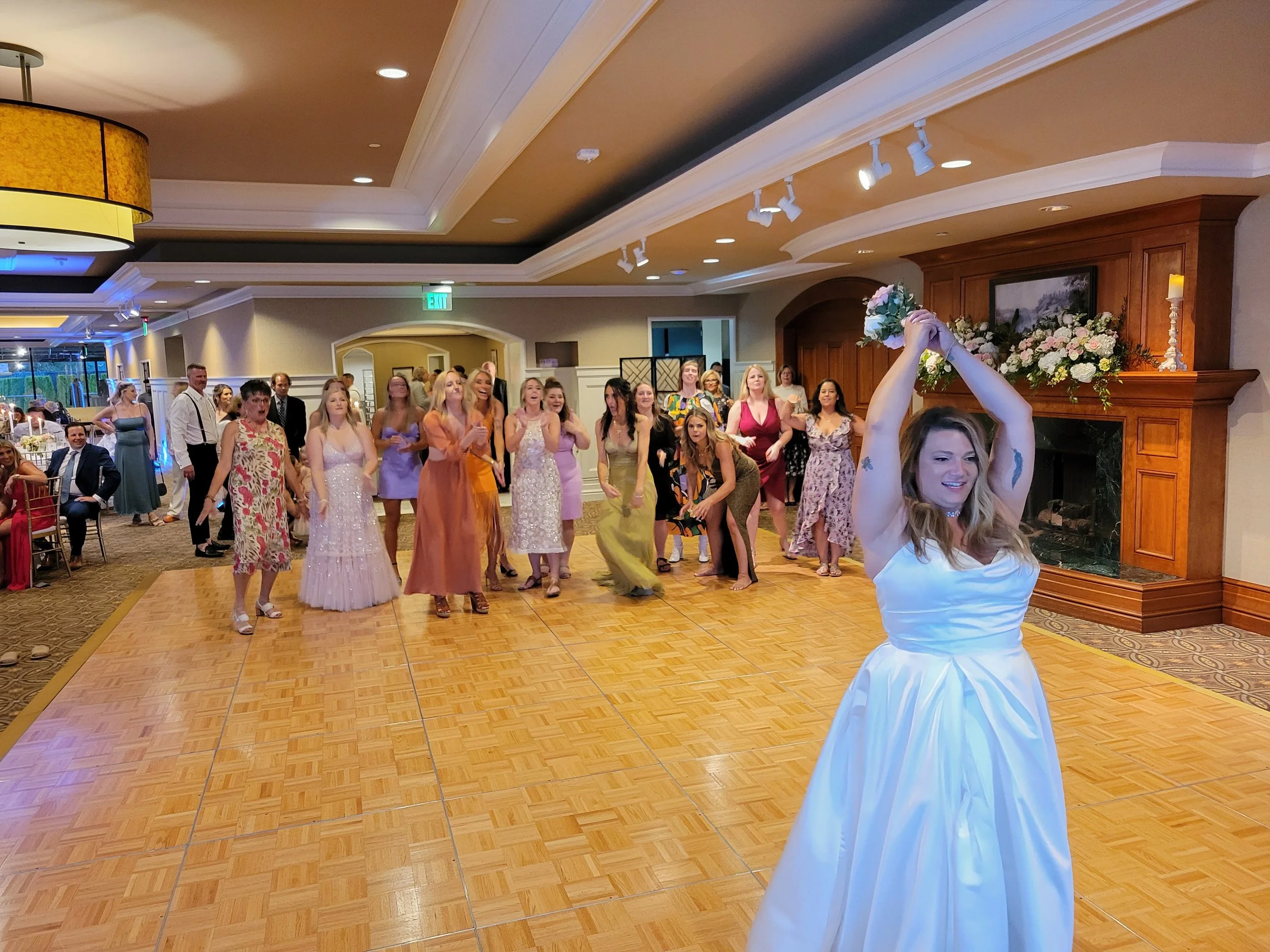 A bride in a white wedding dress throws her bouquet over her head to a group of women standing behind her, ready to catch it, at a wedding reception in a decorated banquet hall.