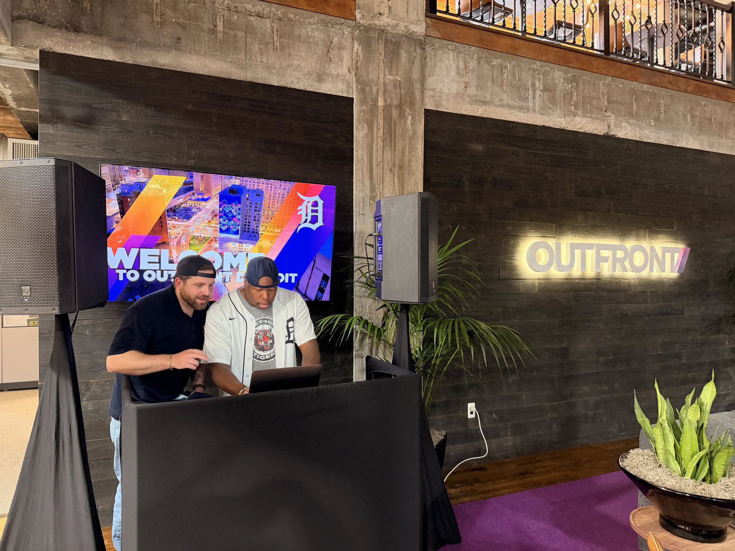 Two men are standing behind a table with a laptop, looking at it together. They are in an indoor space with a dark wooden wall that has a neon sign reading 'OUTFRONT'. A large screen behind them displays a colorful cityscape with the text 'WELCOME TO