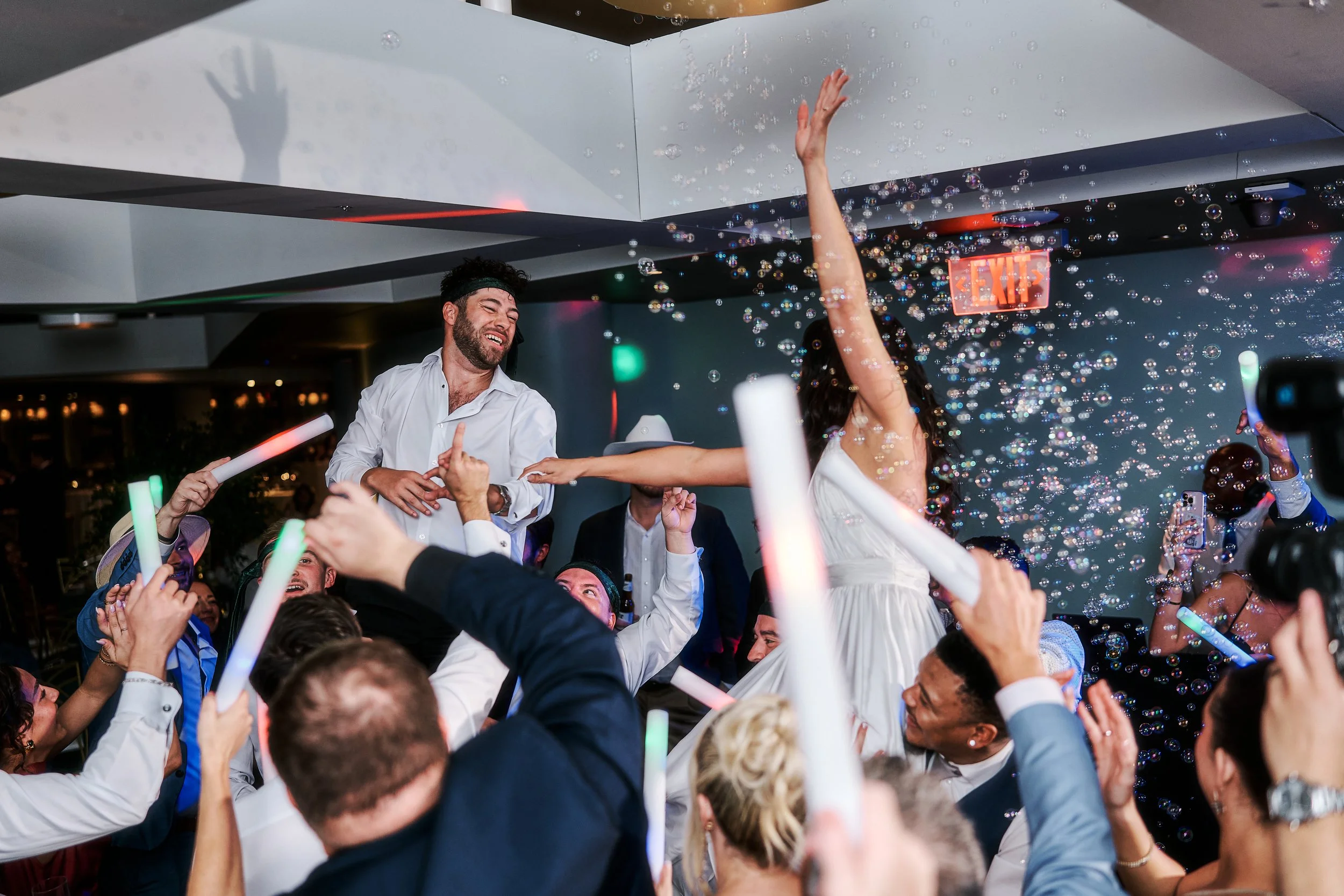 People celebrating at a wedding party with bubbles and glow sticks, with a man and woman being lifted, dancing and enjoying the moment.