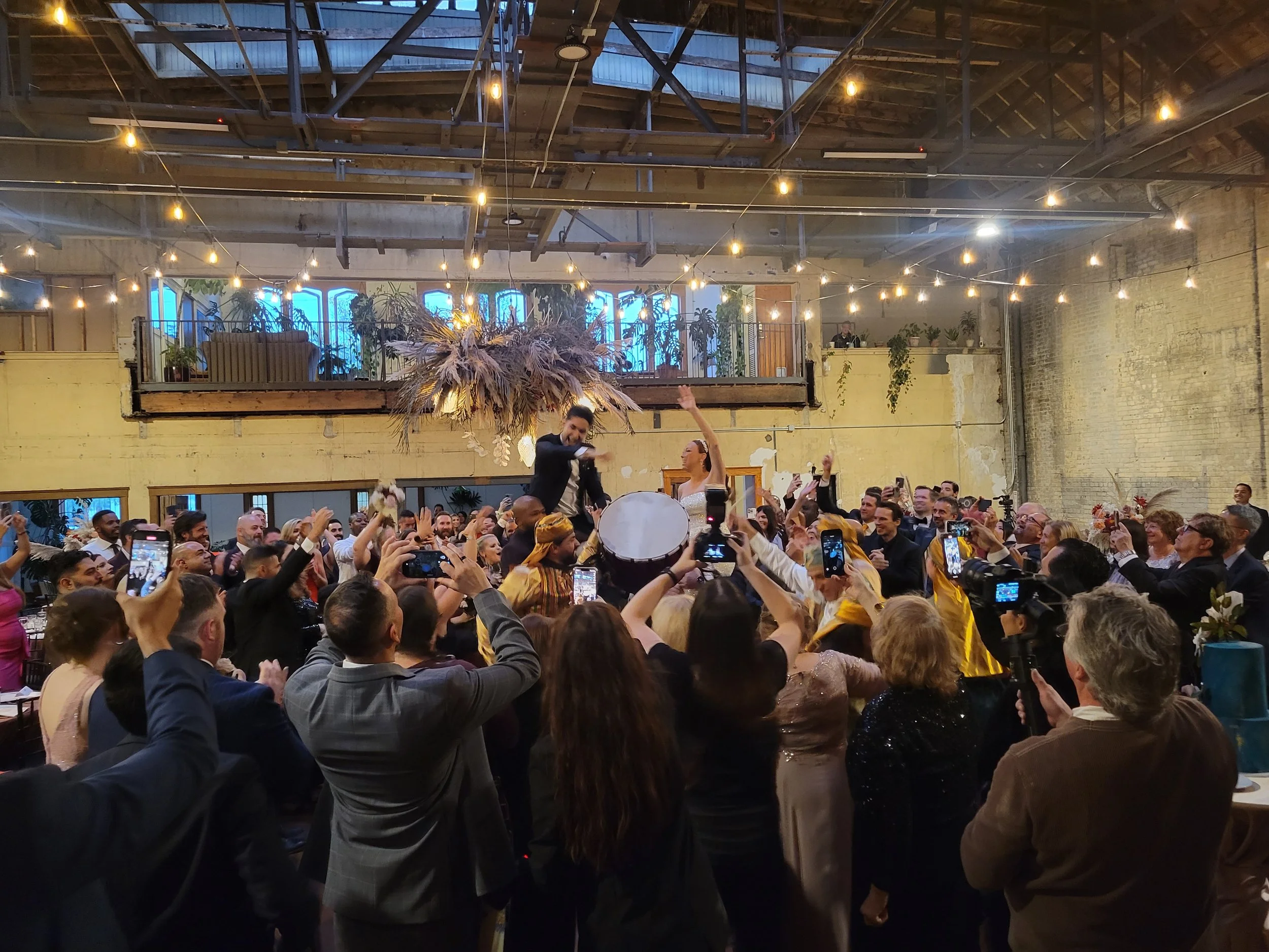 Crowd of people celebrating at a wedding reception with musicians and dancers in an industrial-style venue with string lights and decorative hanging plants.