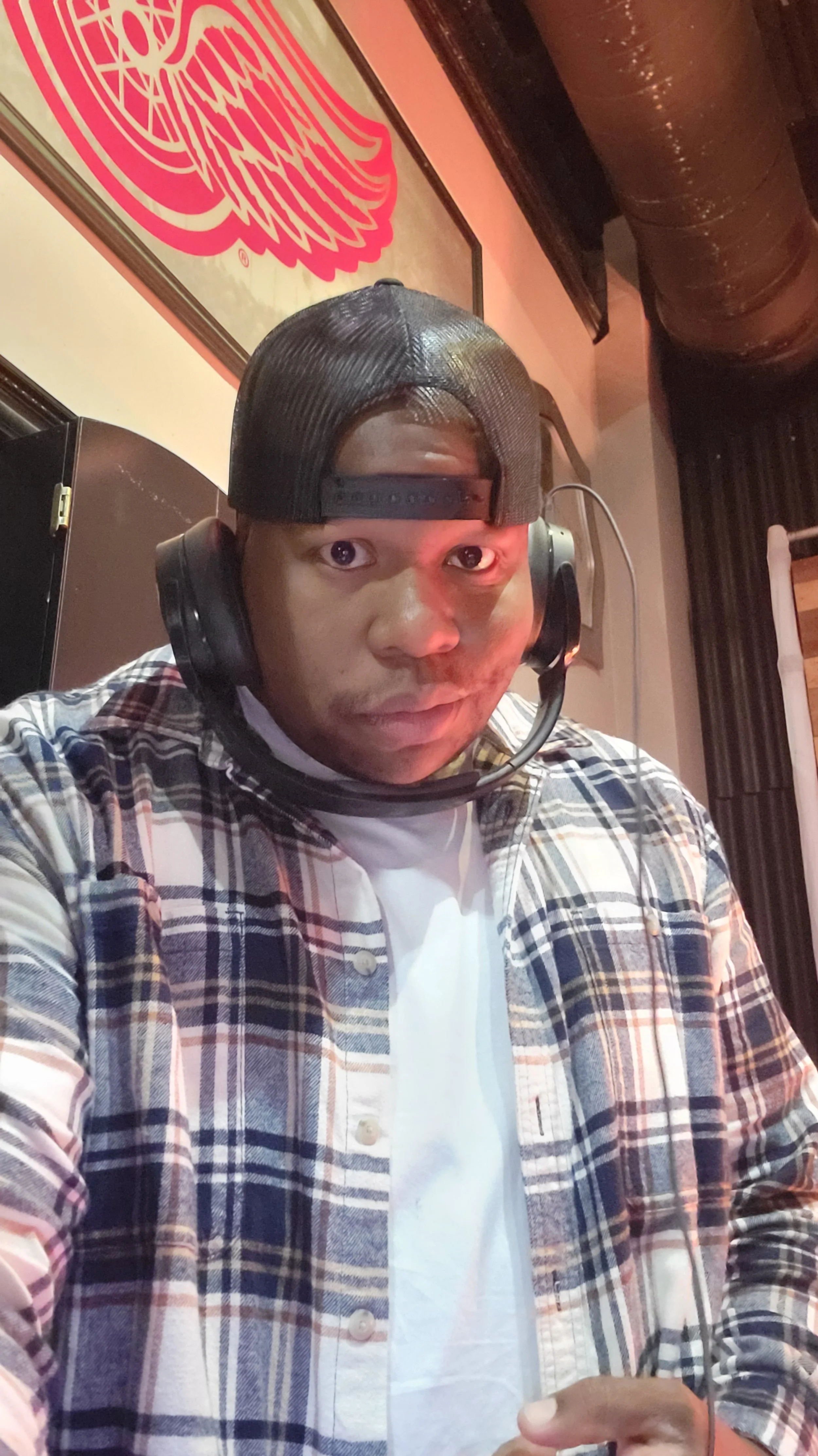A man wearing a black baseball cap backwards, a plaid shirt, and headphones with a microphone. He is looking at the camera with a serious expression. Behind him, there is a wall with a red and white logo or artwork and black curtains.