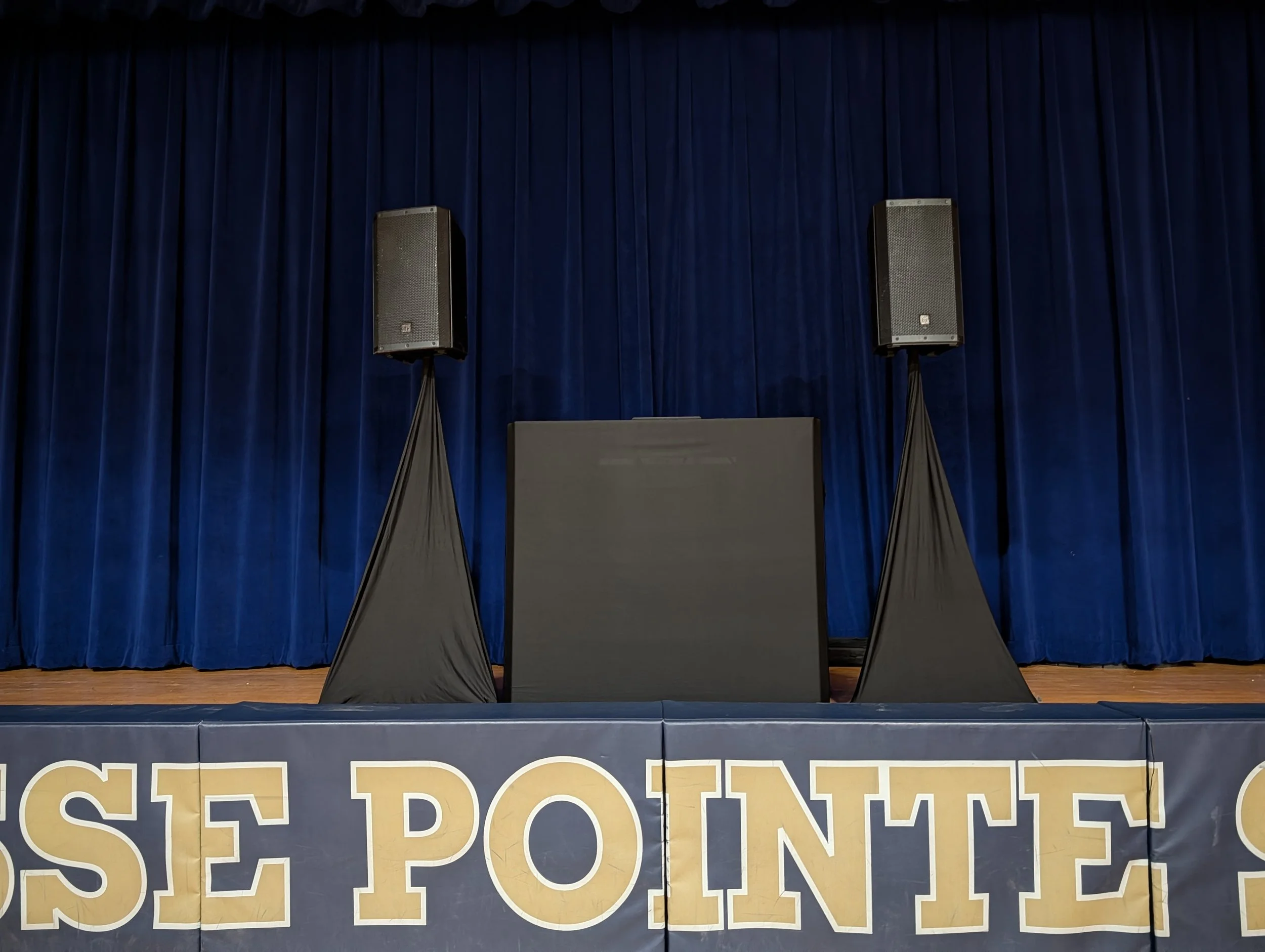 Stage with a blue curtain backdrop, two large speakers on stands, and a black podium in the center. Part of a banner with the words "USE POINT" visible at the bottom.
