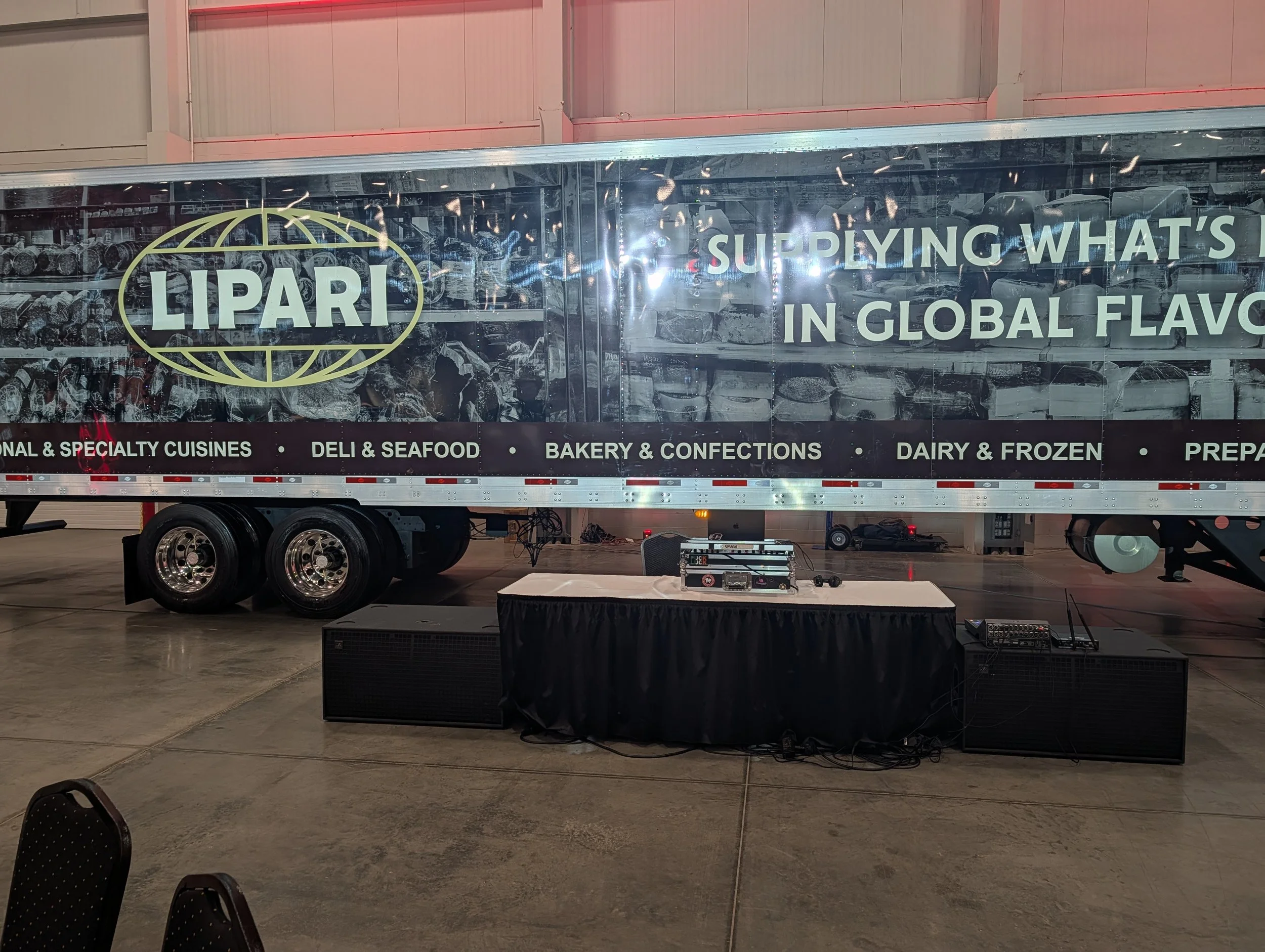 A large warehouse trailer with the LAPARI logo and the phrase 'SUPPLYING WHAT'S IN GLOBAL FLAVOR' written on its side, set up indoors with speaker equipment and a DJ table underneath.