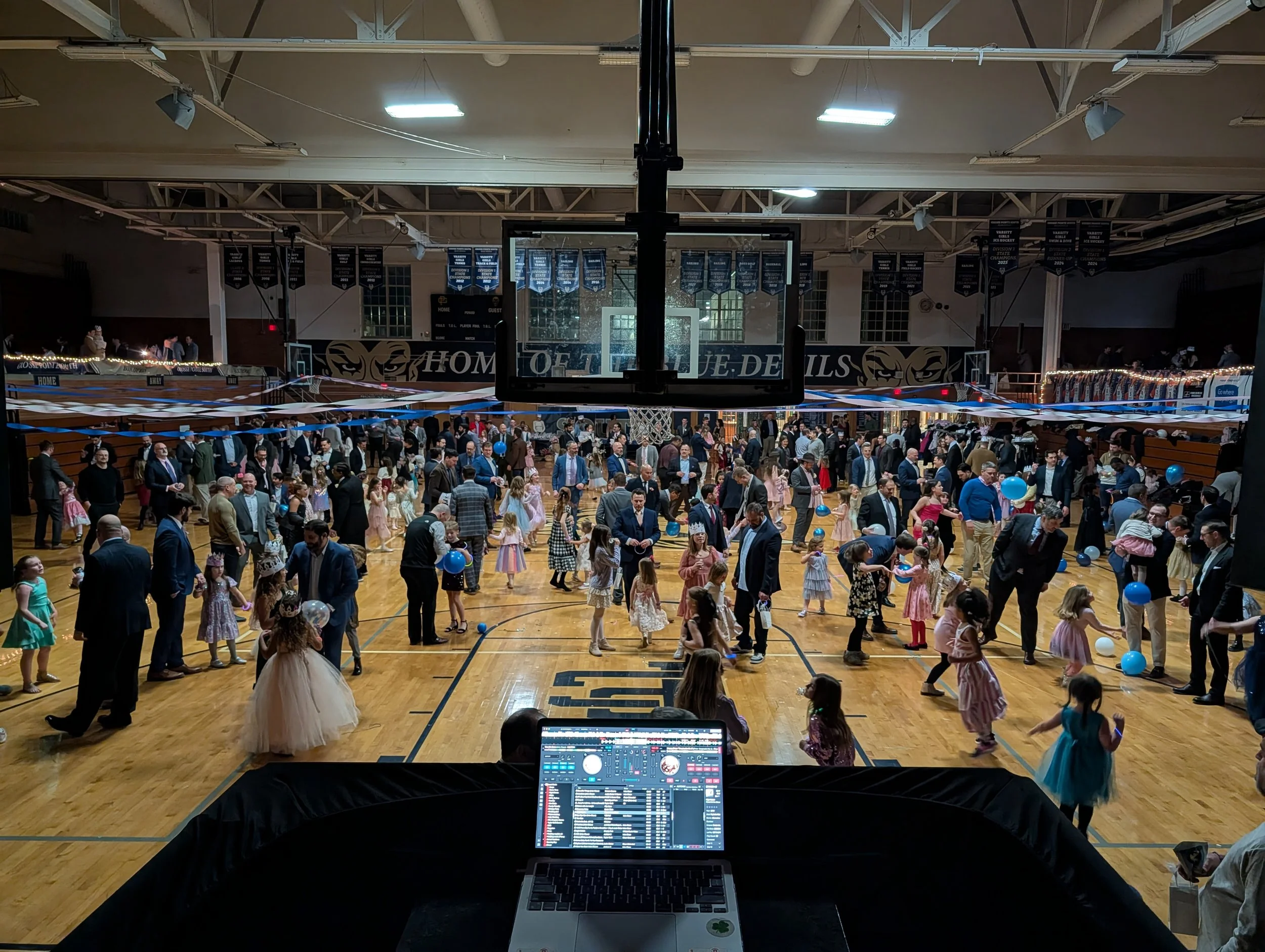 A gymnasium decorated for a dance party with children and adults dancing on the floor. Balloons and ribbons decorate the gym. A DJ booth and speakers are in the foreground.