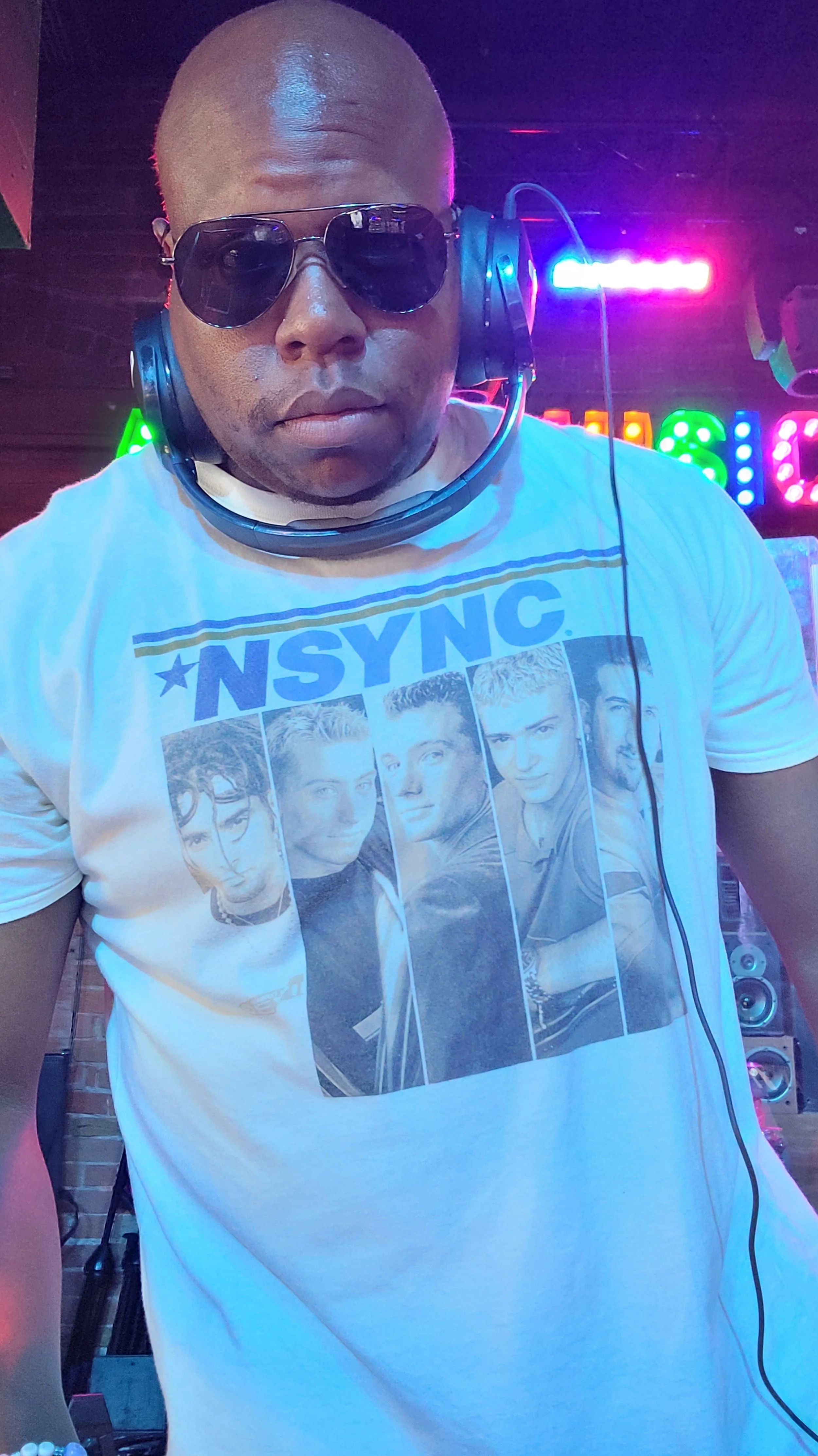 A man wearing sunglasses and a headphone around his neck, standing in a colorful, vibrant environment, possibly a club or music venue.