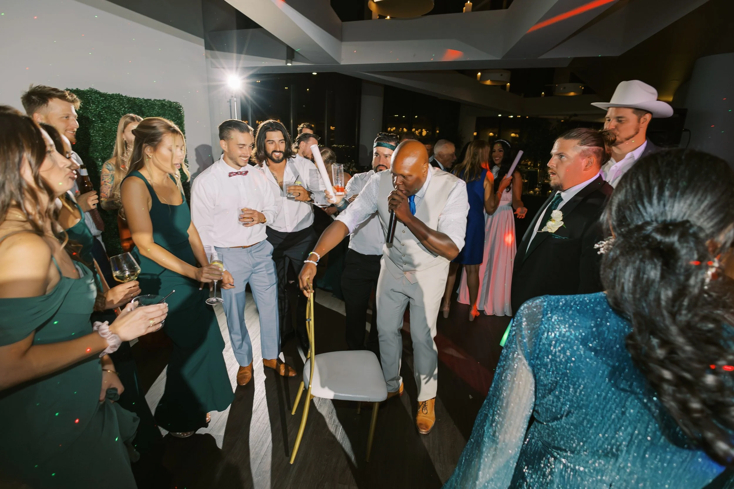 People gathered at a celebration, with a DJ or performer in the center, holding a microphone and a cane, surrounded by party guests in formal attire, in a modern indoor venue with dim lighting and colorful spotlights.