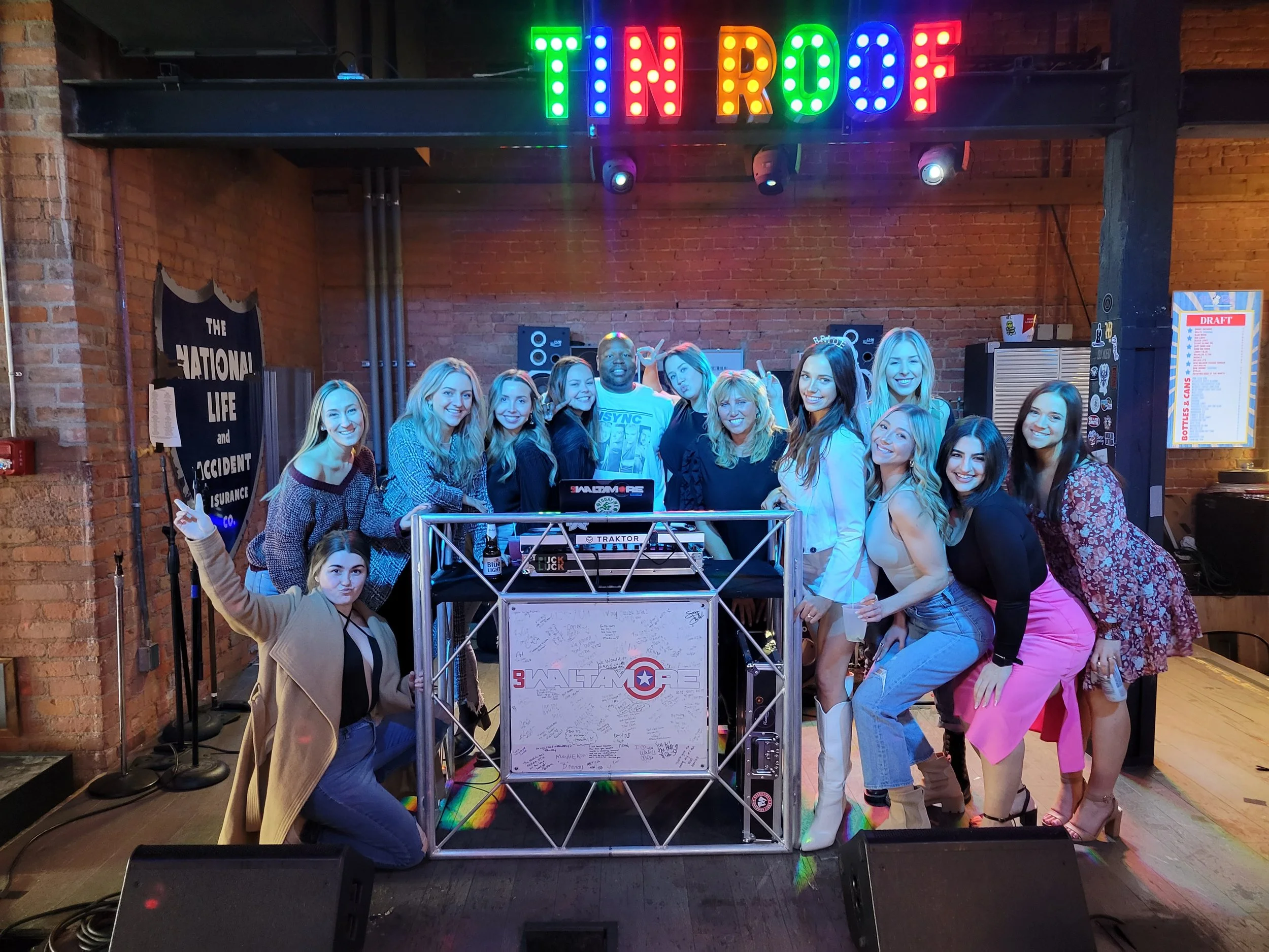 Group of people, mostly women, smiling and posing in a venue with a DJ booth labeled "WALTMARE" and a colorful sign above that says "TIN ROOF." Brick walls and stage lighting are visible in the background.