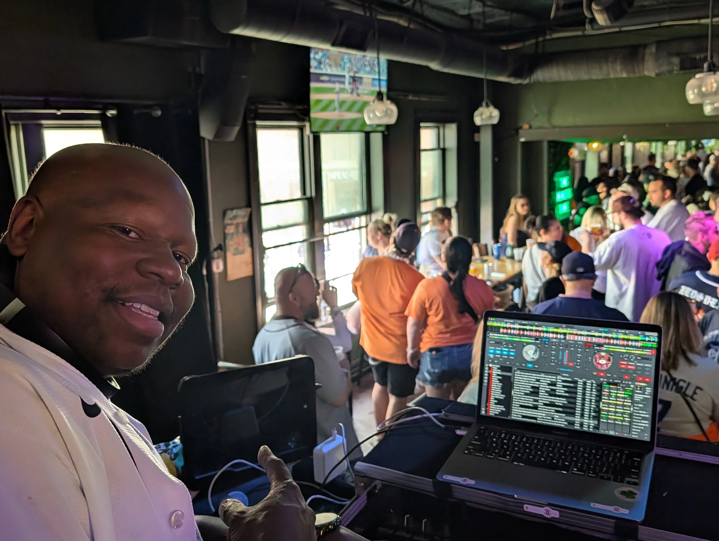 A man smiling and pointing at a laptop with DJ equipment in a crowded indoor venue enjoying a live event.