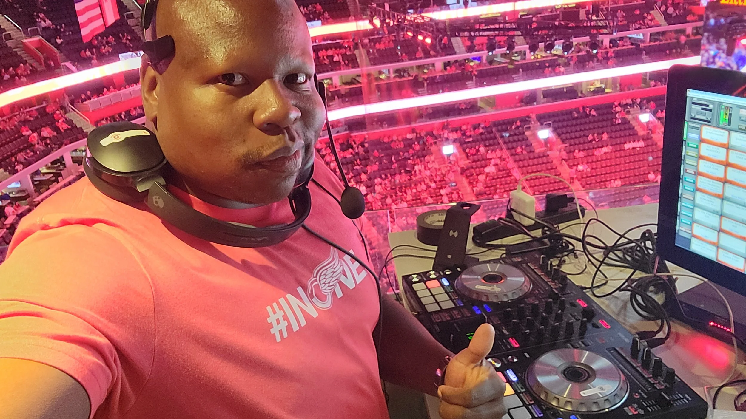 DJ wearing a pink t-shirt, headphones, and speaking into a mic at a concert, with a large audience in a stadium with pink lighting in the background.