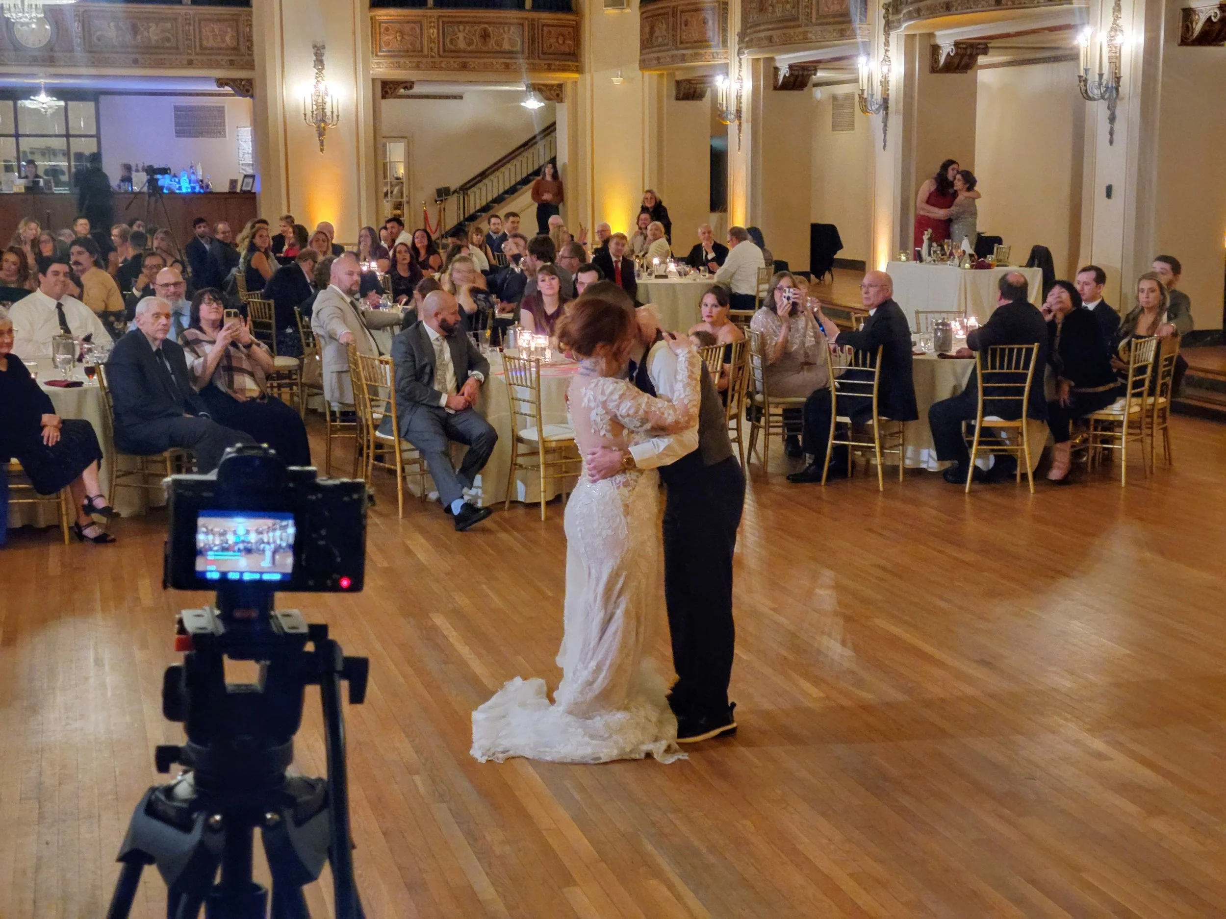 A bride and groom sharing their first dance at a wedding reception inside a banquet hall, with guests seated at round tables watching and recording the moment.