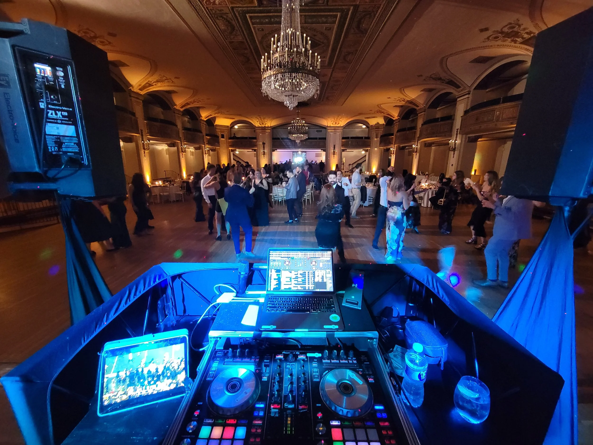 View from a DJ's booth overlooking a dance floor in an elegant ballroom with chandeliers and ornate ceiling decorations, filled with people dancing.