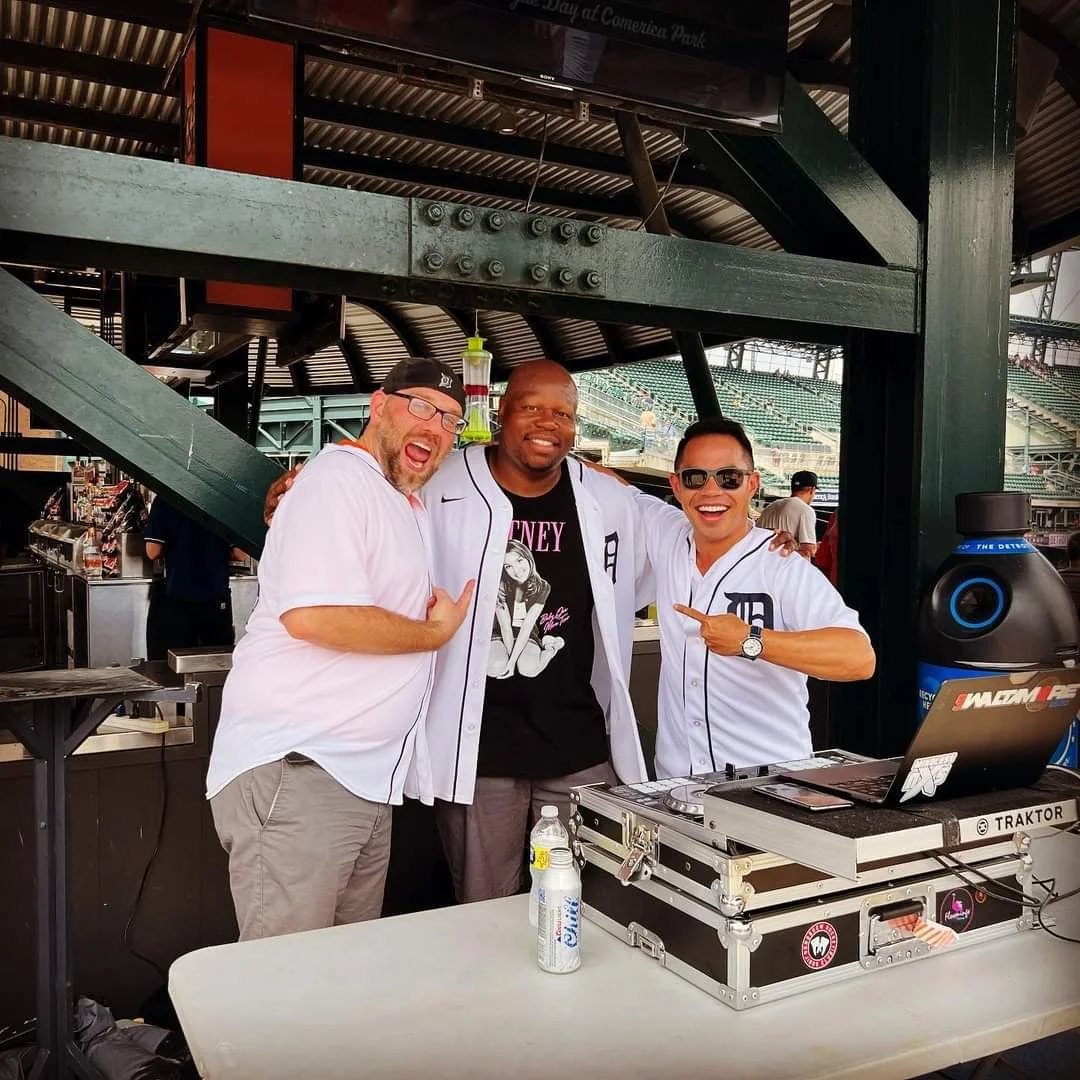 Three men standing behind a DJ table at Comerica Park, smiling and celebrating, with stadium seating in the background.