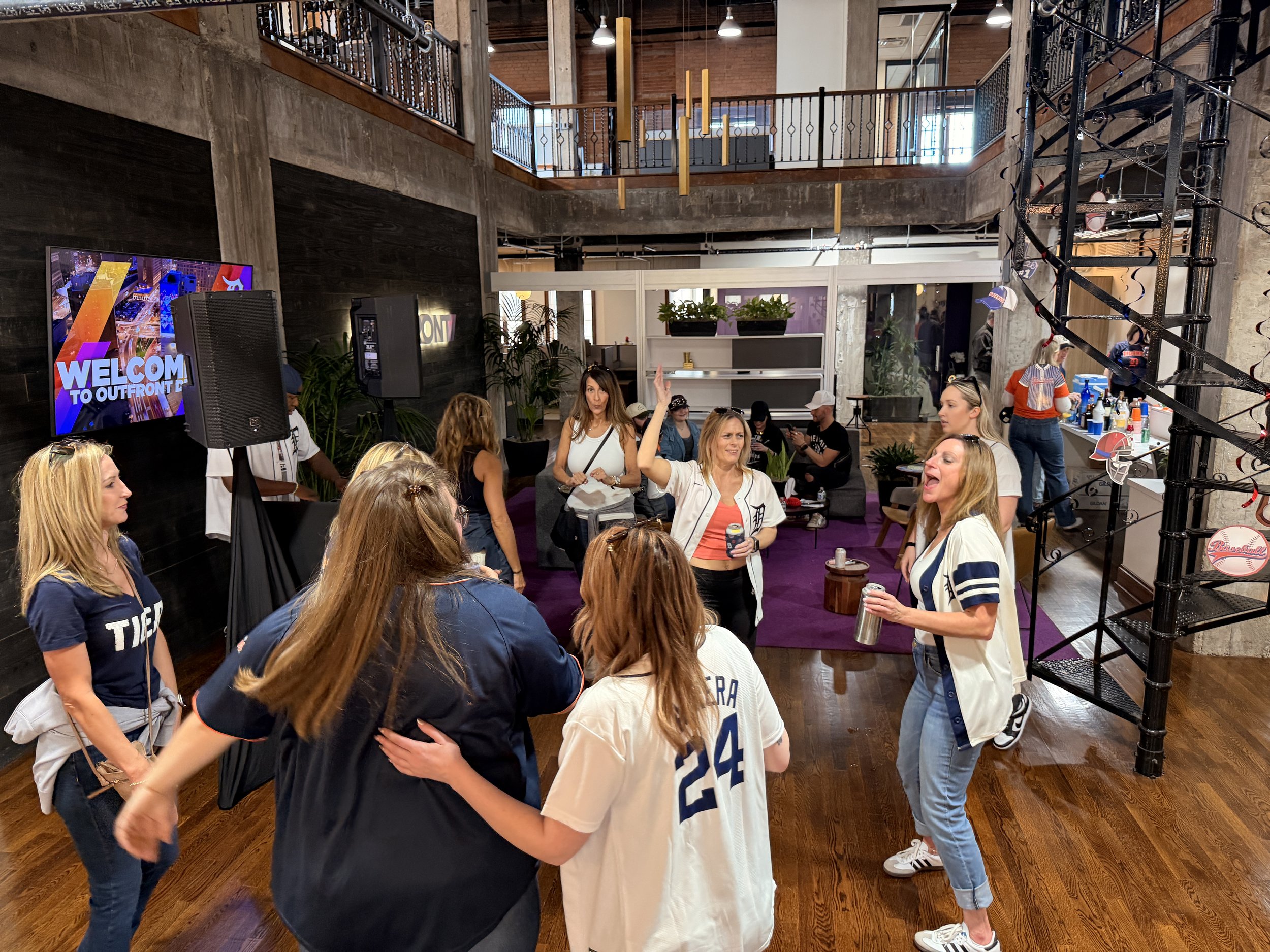 People gathered at a social event with some dancing and talking, in a modern indoor space with purple carpet, plants, and a large screen displaying 'Welcome to Outfront.'