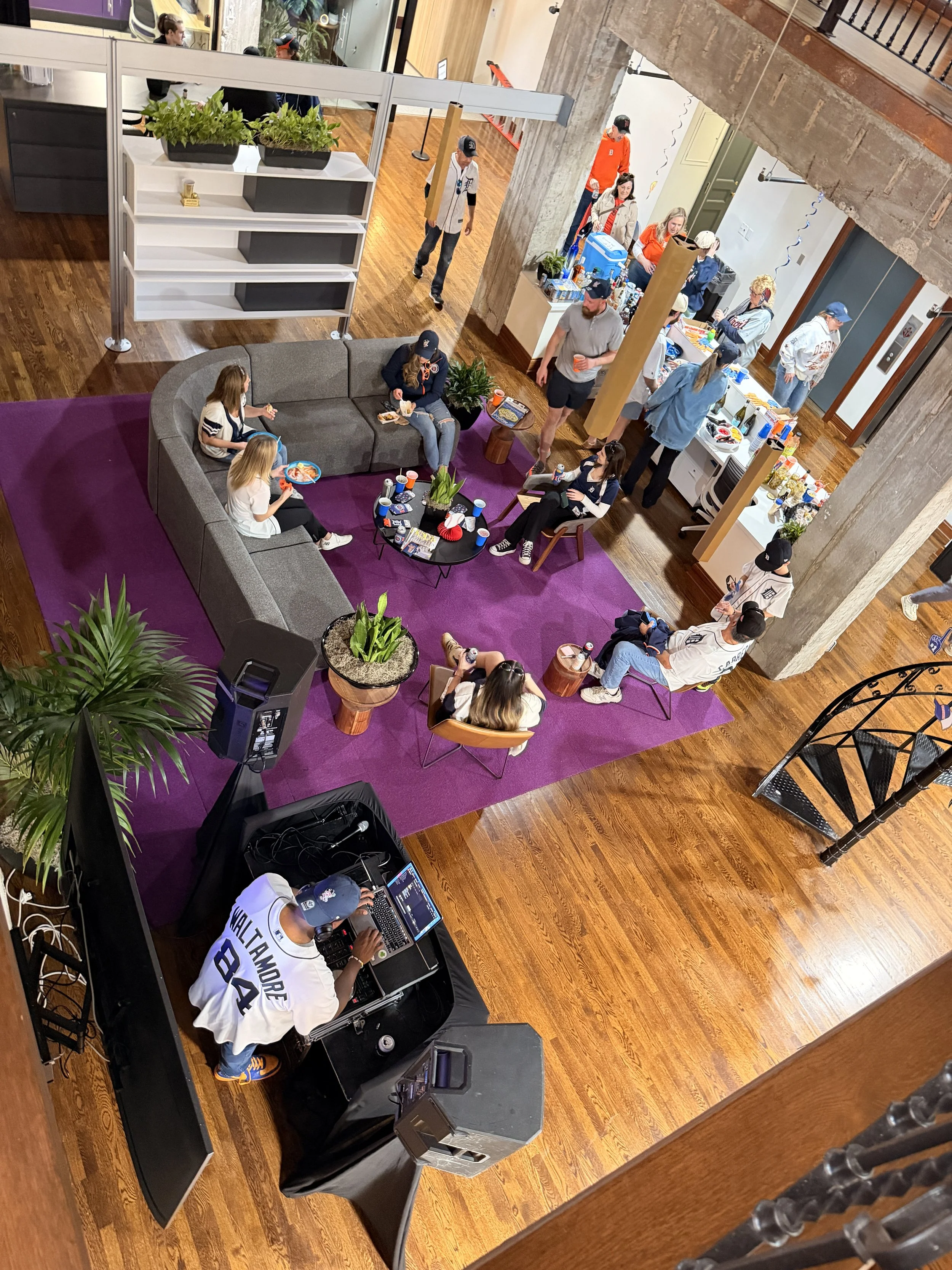 People gathering at a social event in a spacious venue, with a DJ playing music, a lounge area with a purple rug and a sofa, and food and drinks on a table near the counter.