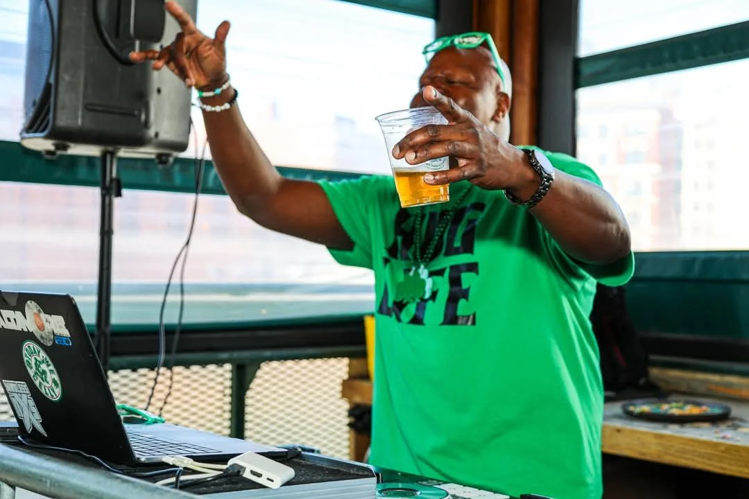 A man wearing a green t-shirt and sunglasses dancing and holding a drink while DJ equipment is nearby.