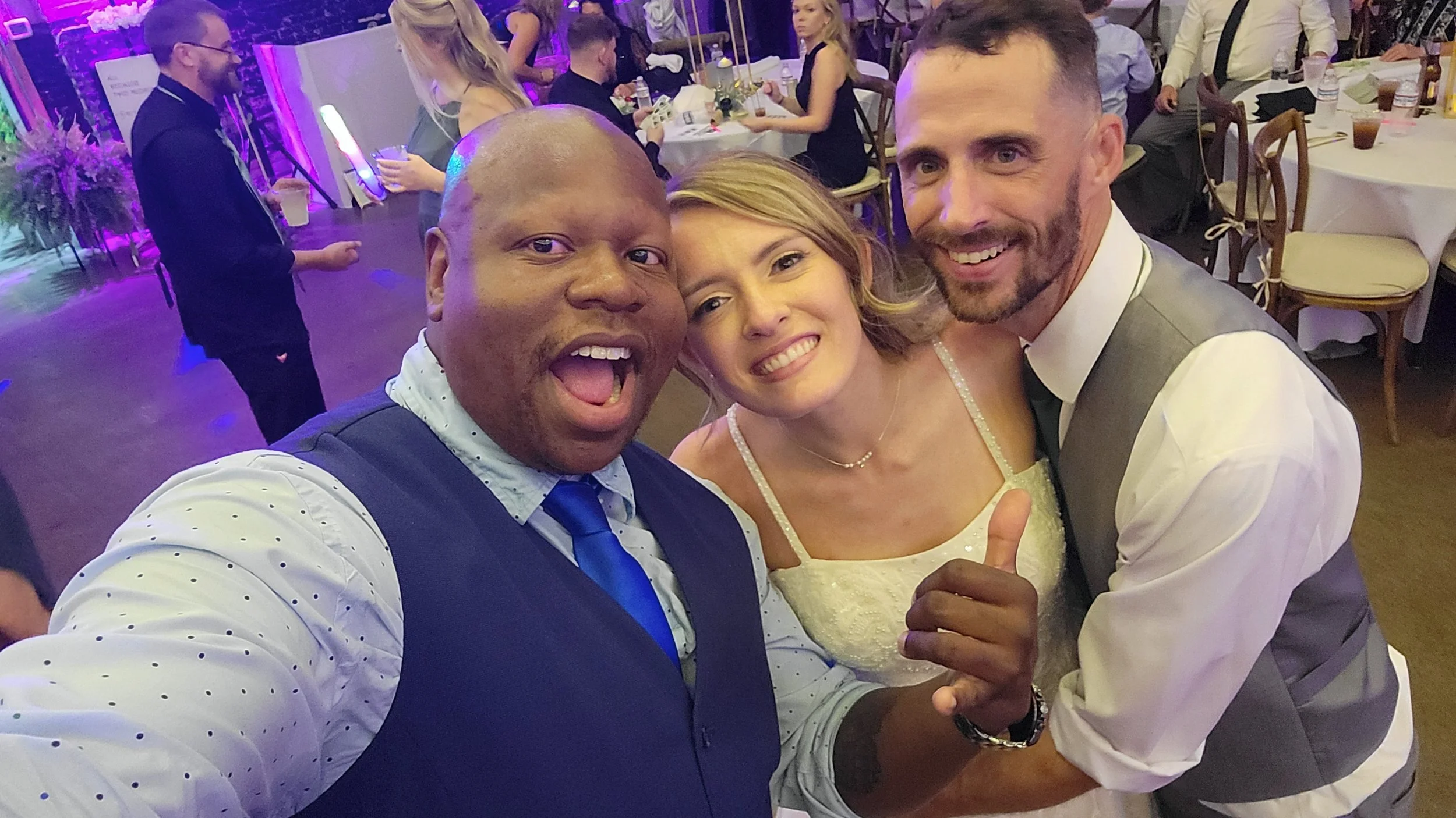 Group of three friends smiling and taking a selfie at a wedding reception. The woman is wearing a white dress, and the two men are in formal attire. The background shows other guests seated at round tables and a dance floor with purple lighting.