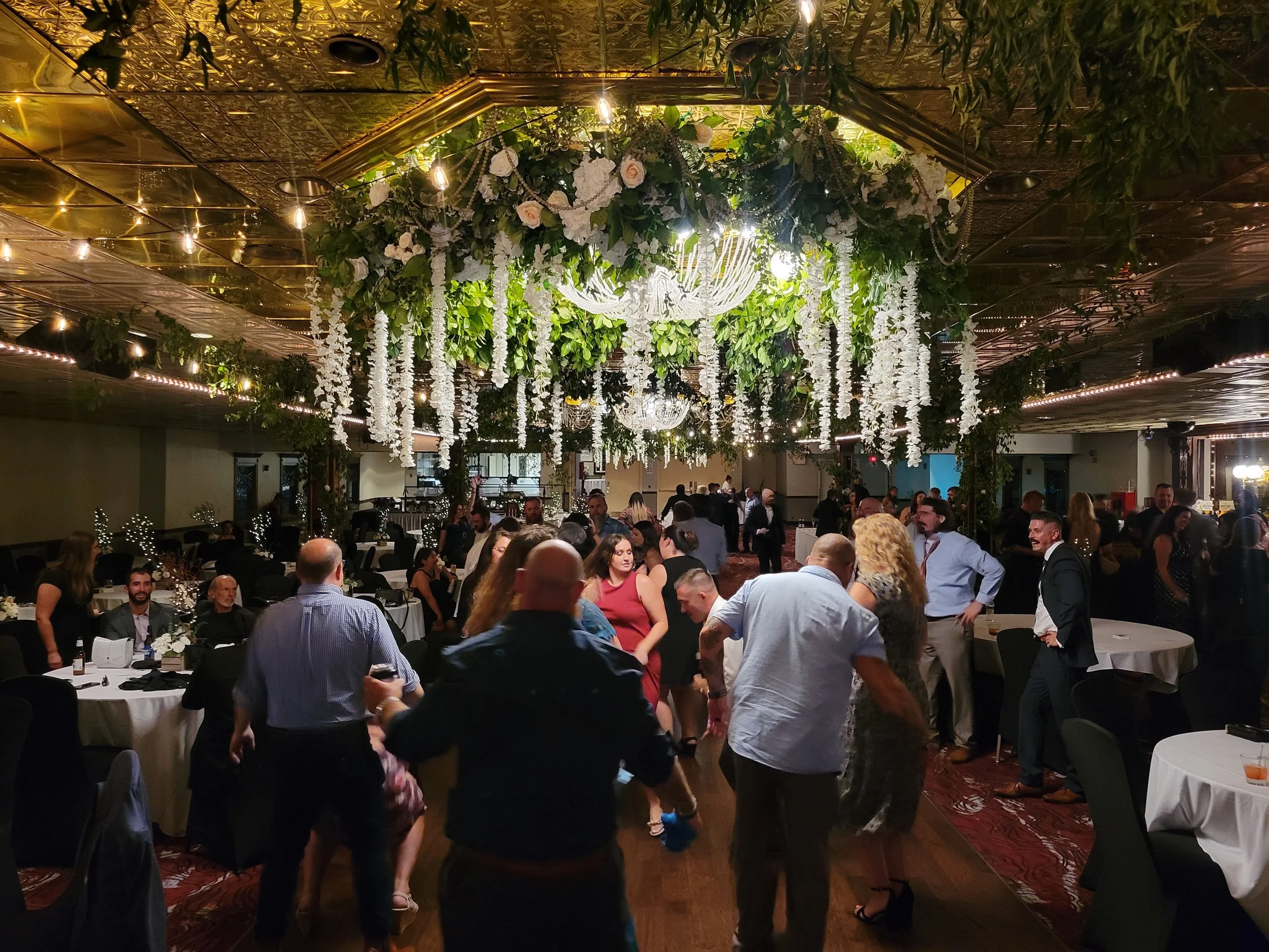 People dancing and socializing at a banquet hall decorated with hanging white flowers, greenery, and chandeliers.