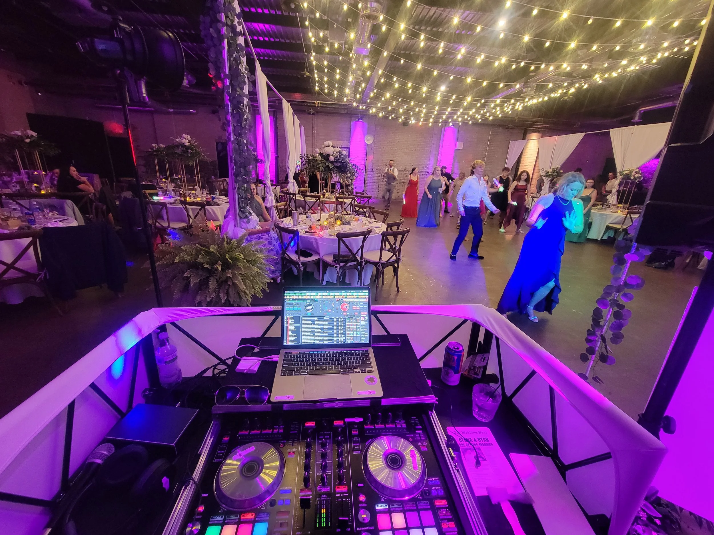 View from DJ booth overlooking a dance floor at a party or wedding reception. The dance floor has people dancing, and decorated tables with floral centerpieces. Ceiling is adorned with string lights, creating a festive atmosphere with purple and yell
