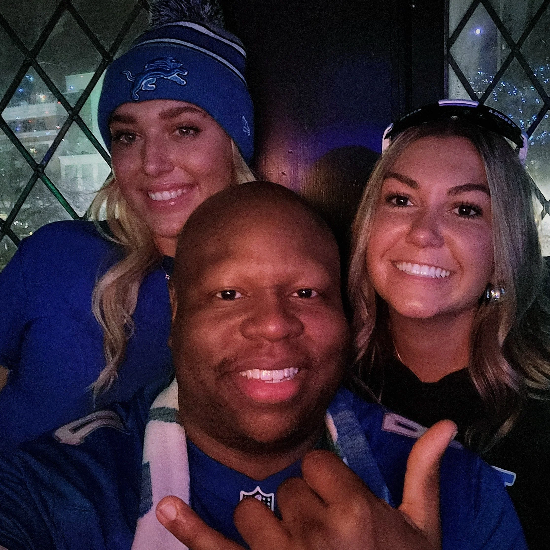 Three smiling people, two women and one man, are close together, with the man in front. The woman on the left is wearing a blue beanie and a sweat shirt with a logo, and the woman on the right has long dark hair and is wearing sunglasses on her head.