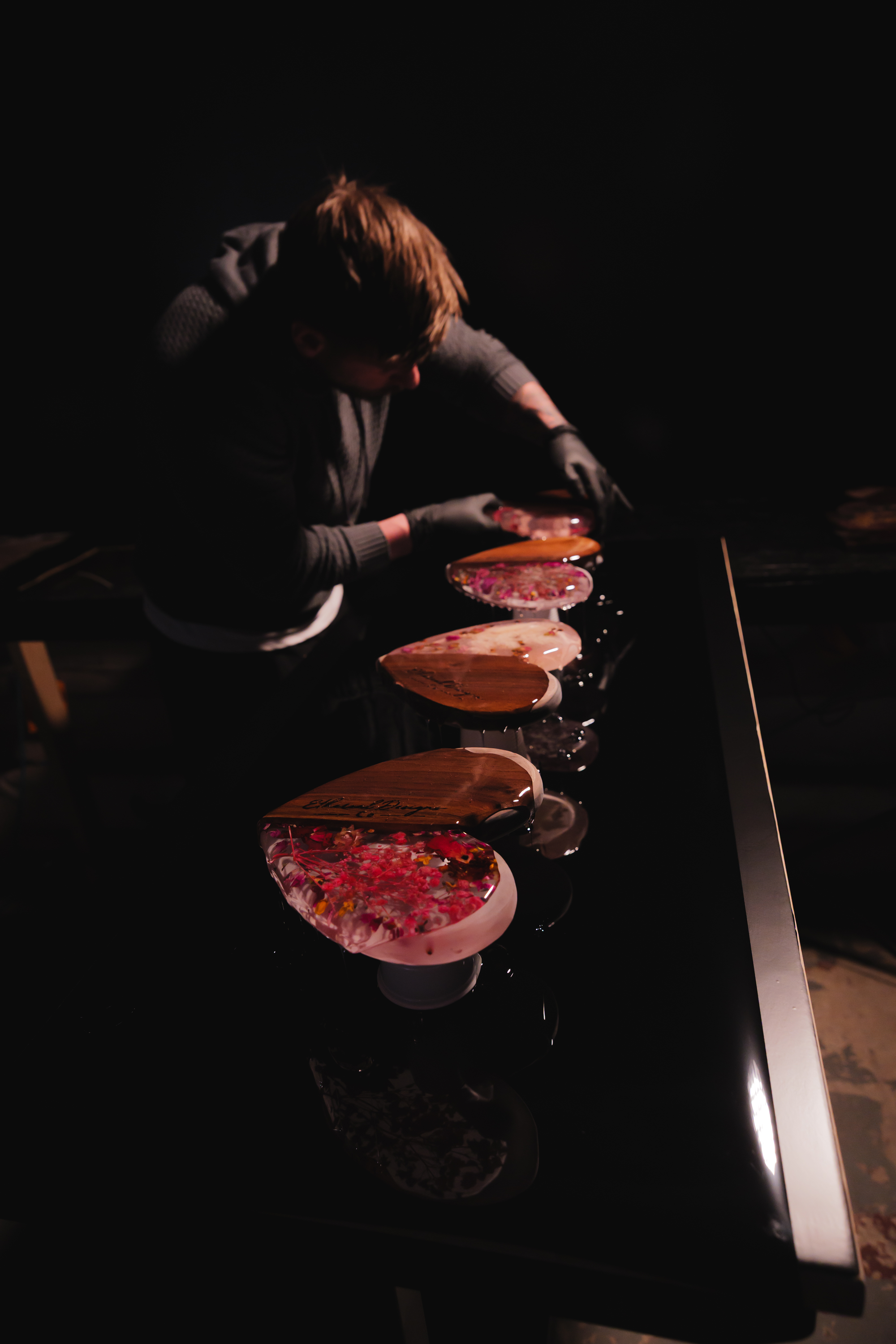 A person with brown hair wearing gray shirt and black gloves is arranging decorated heart-shaped objects on a black table. The objects appear to be made of resin with floral patterns inside. The scene is dimly lit with focused light on the objects.