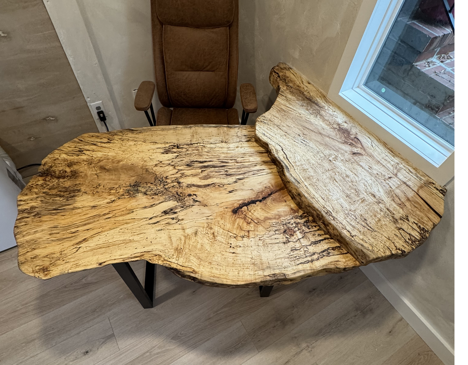 Close-up of a rustic, live-edge wooden table with a natural, irregular shape placed in a room with a light-colored wall and a large window. A brown office chair is positioned behind the table.
