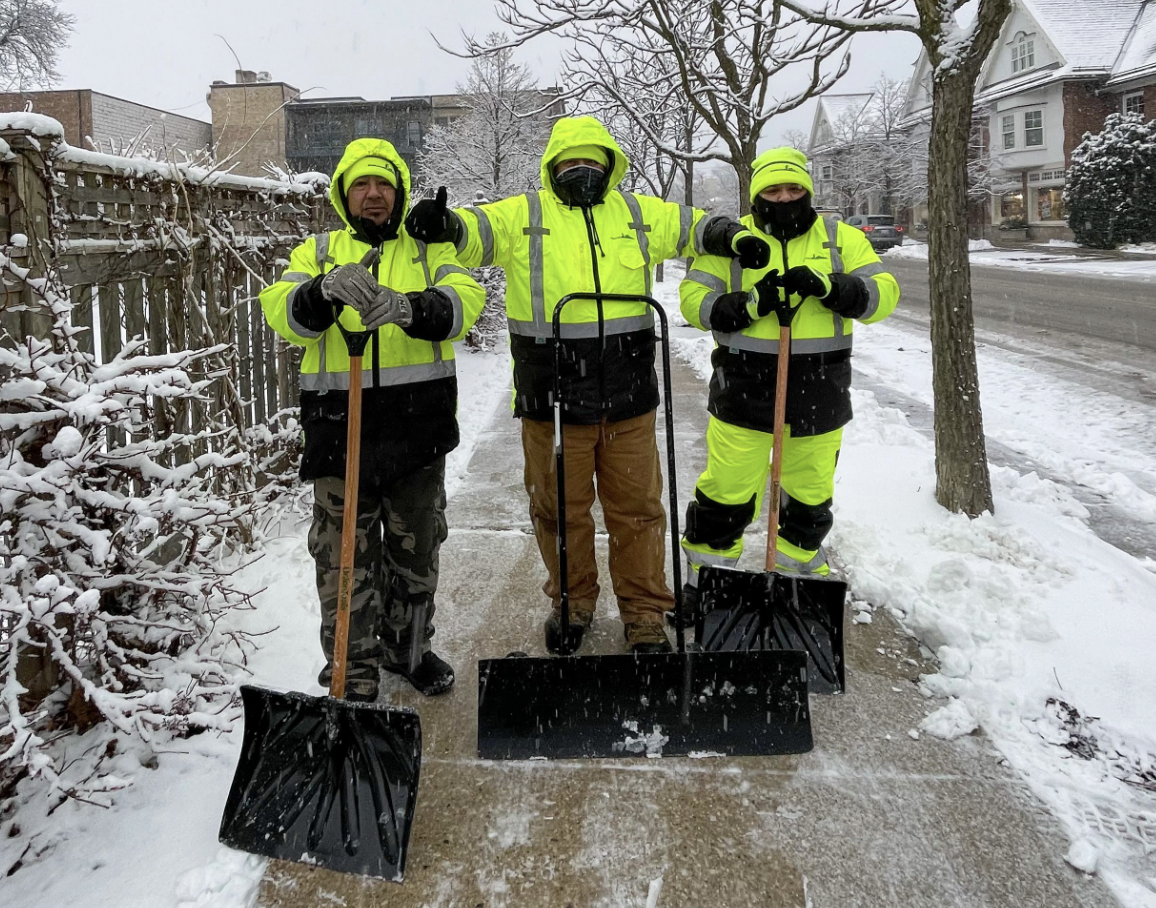 Three workers in high-visibility jackets and hats clearing snow from a sidewalk with snow shovels during winter.