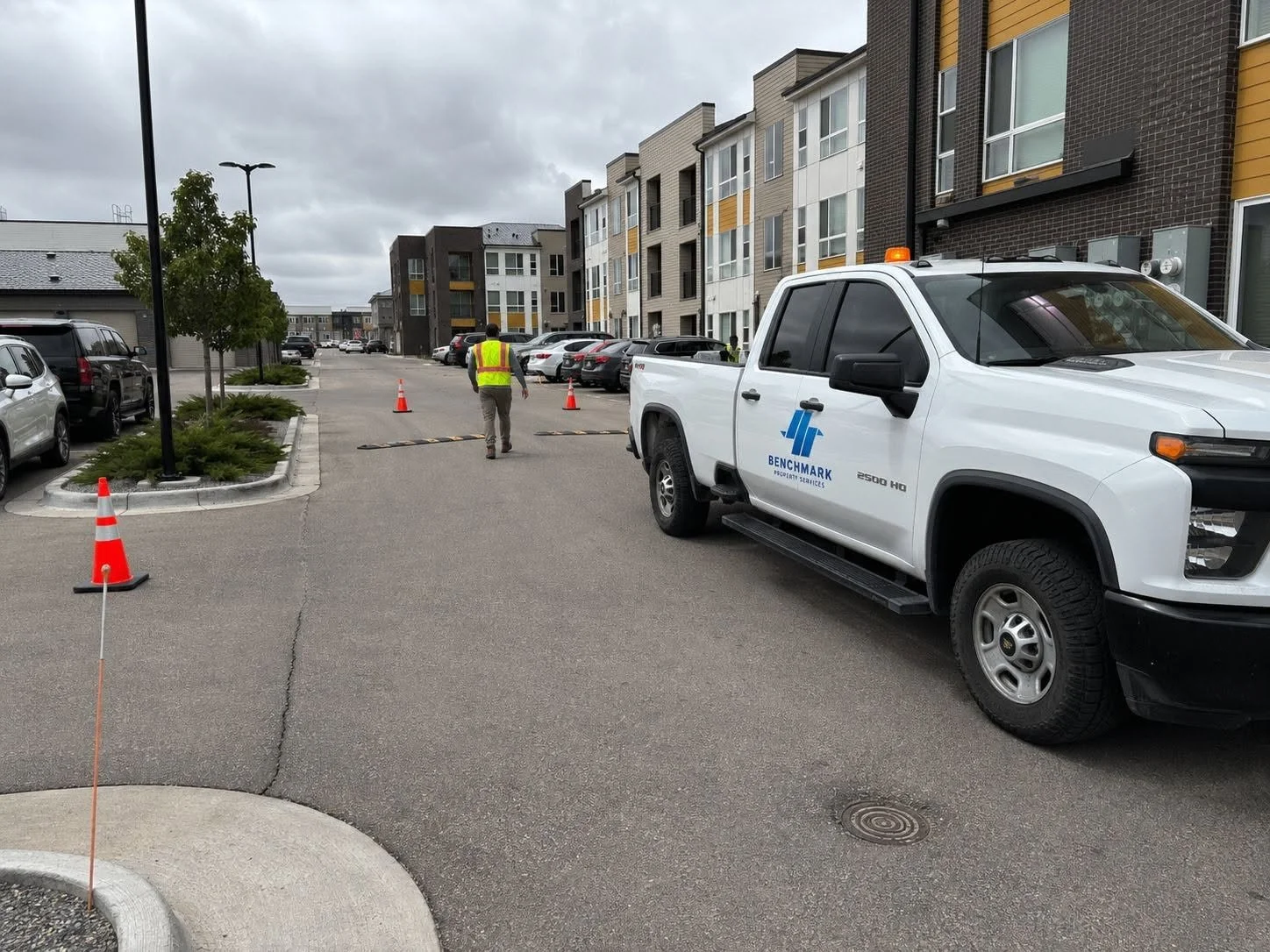 A parking lot with multiple parked cars, a man in a high-visibility vest walking towards the back, a white truck with a blue logo and safety lights on top, orange cones marking the area, and residential buildings in the background under cloudy skies.