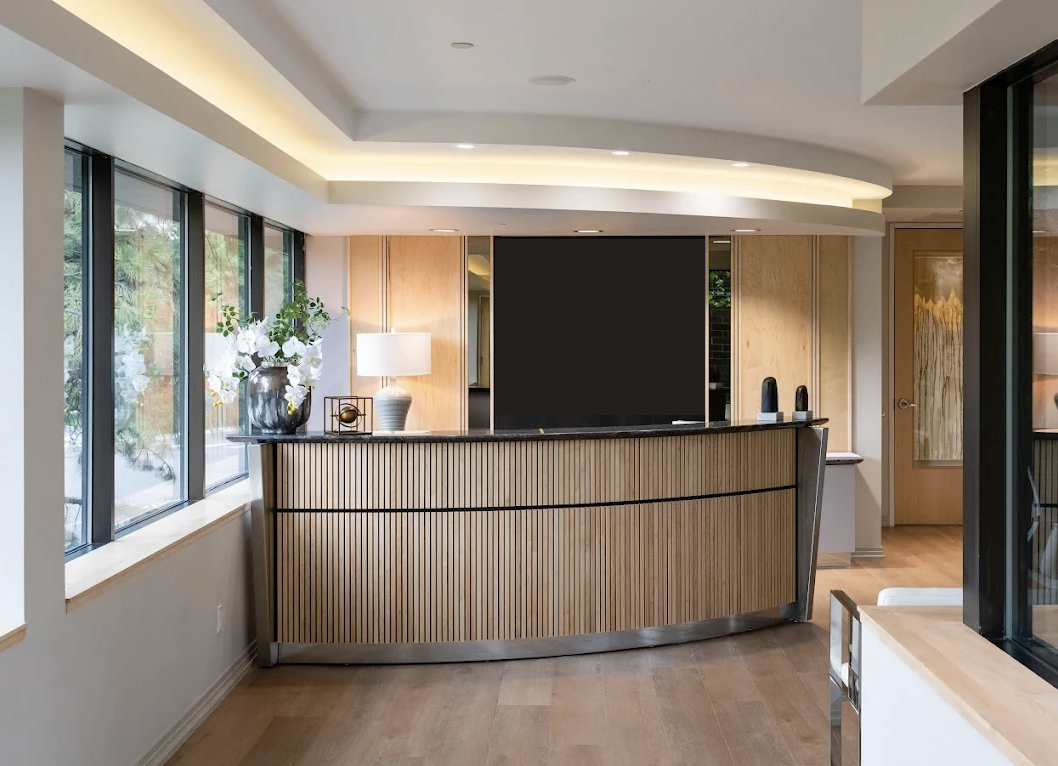 Modern reception desk with a black monitor, white lamp, decorative objects, and a flower arrangement in a sleek, well-lit interior space with large windows and wooden accents.