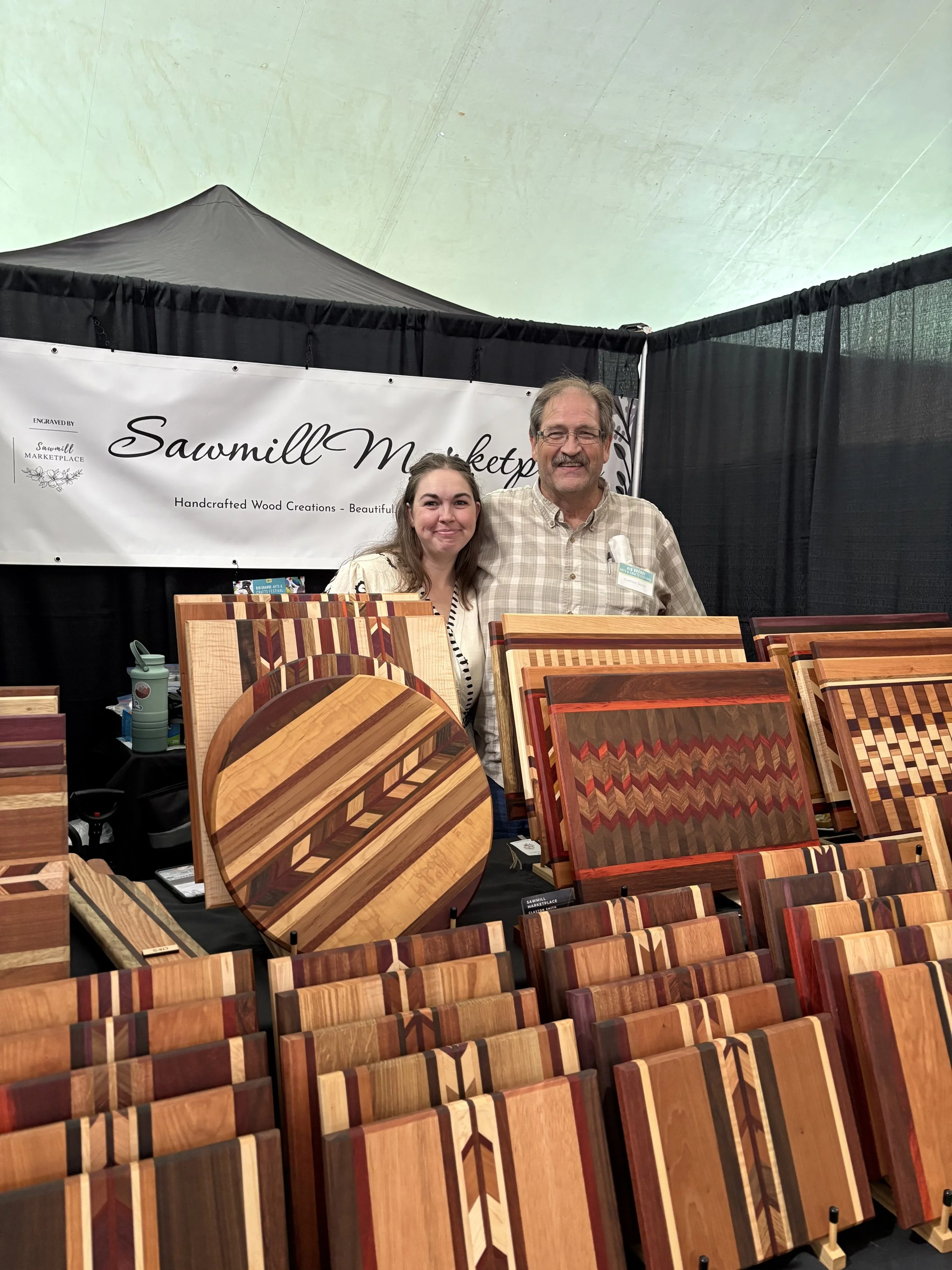 A woman and a man standing behind a table of various handcrafted wood cutting boards at a marketplace booth labeled 'Sawmill Marketplace', with black curtains and a white sign.