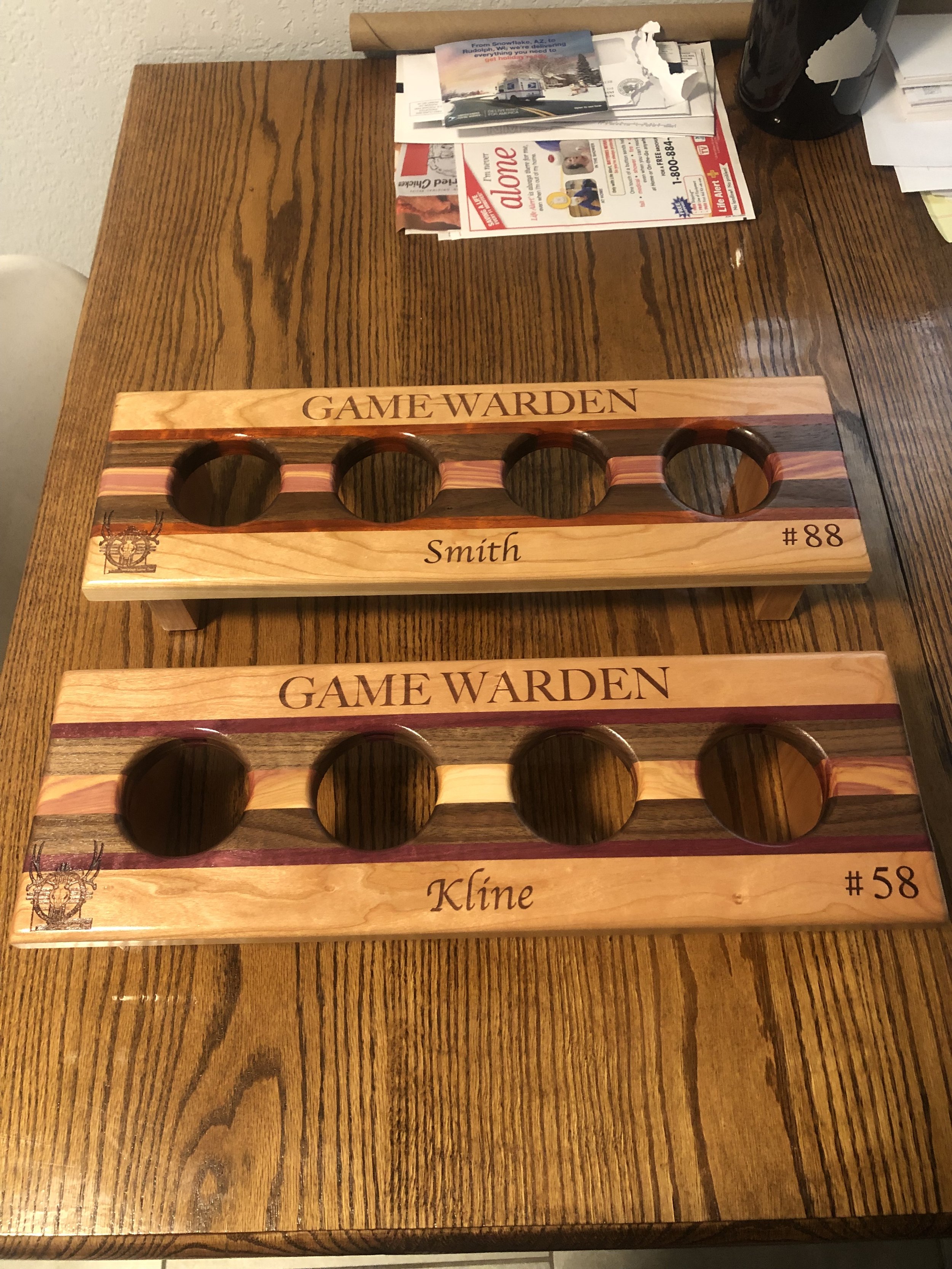 Two wooden game boards labeled 'GAME WARDEN' with holes, one labeled 'Smith' with #88 and the other labeled 'Kline' with #58, on a wooden table. There are papers, magazines, and a black container in the background.