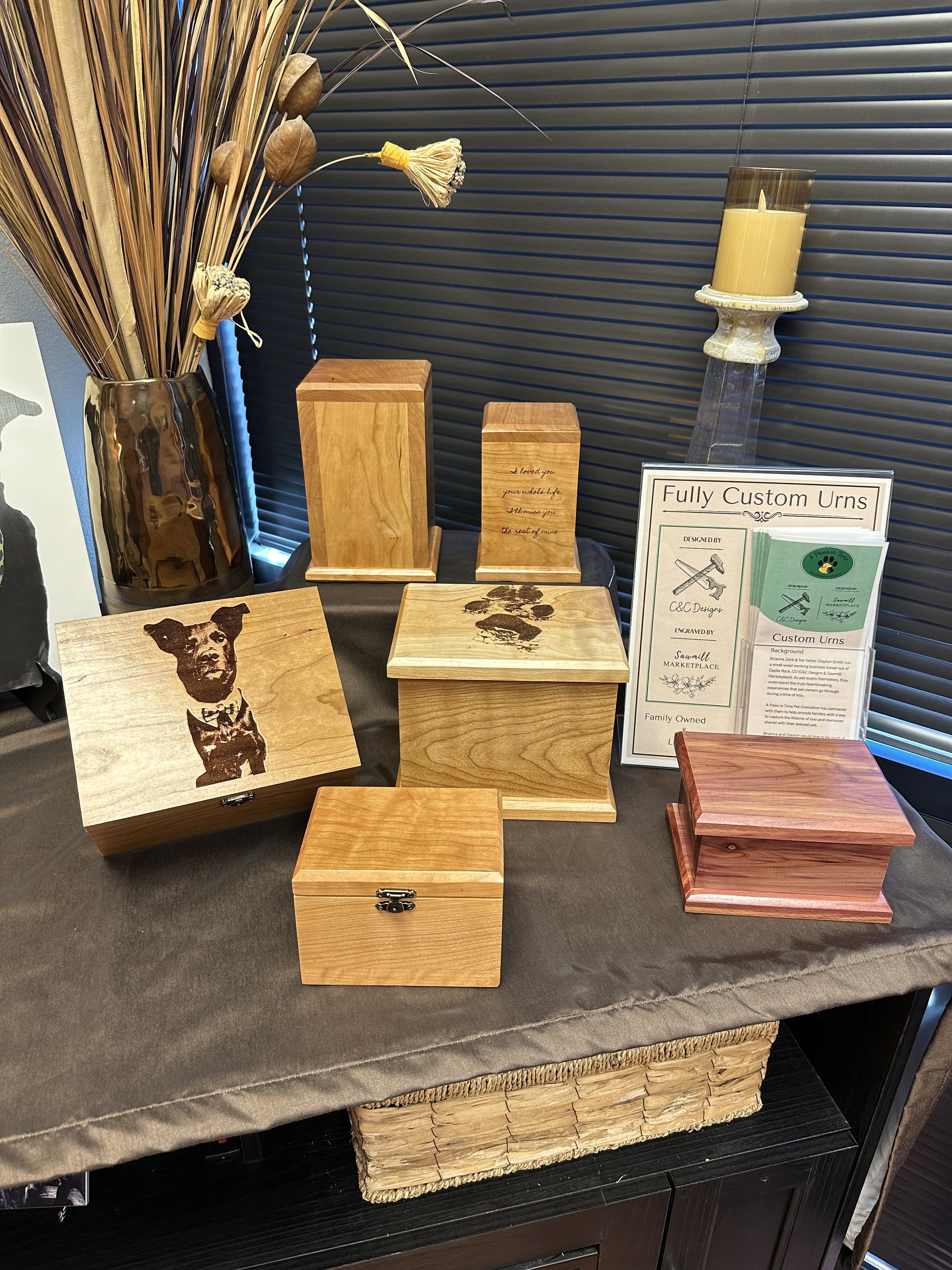 Display of wooden handcrafted urns and decorative items at a marketplace stall, with a sign reading 'Fully Custom Urns' and a framed artwork of a dog on the table.