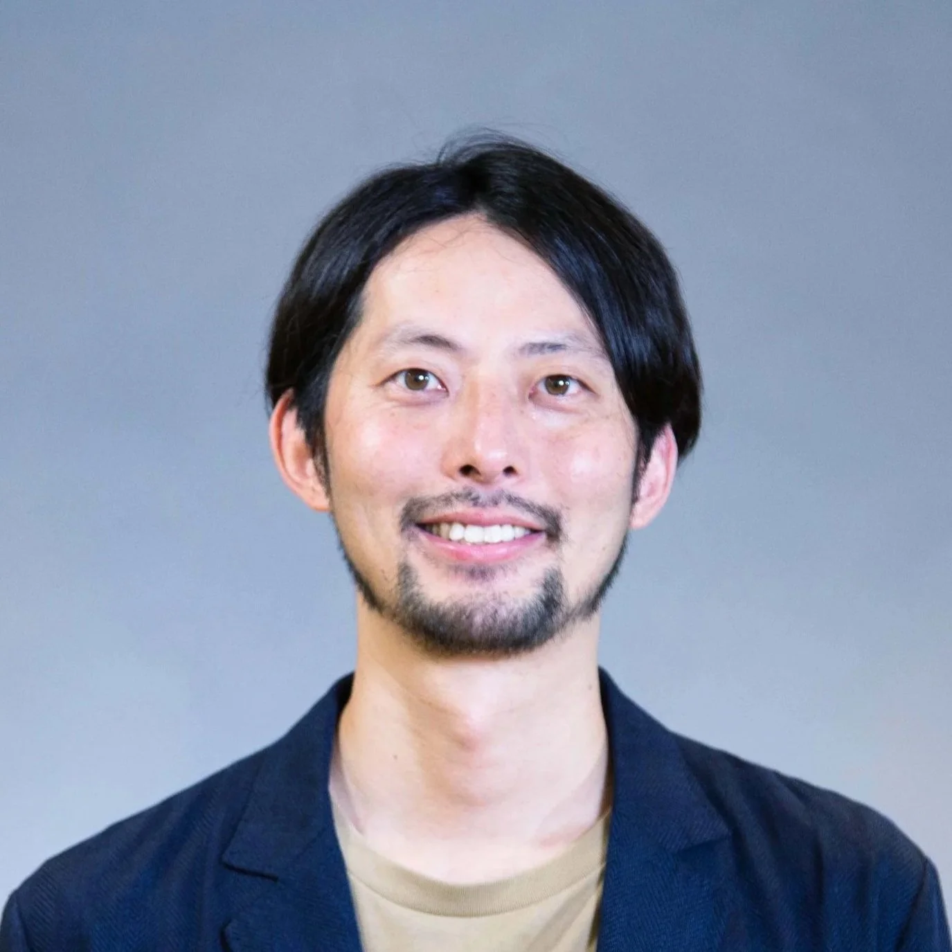 A portrait of a man with black hair, a beard, and mustache, smiling at the camera, wearing a dark blazer and a beige shirt against a neutral background.