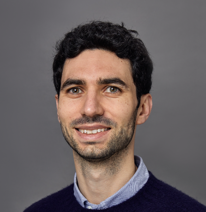 Portrait of a young man with dark curly hair, light skin, blue eyes, smiling, wearing a collared shirt and navy sweater, against a plain gray background.