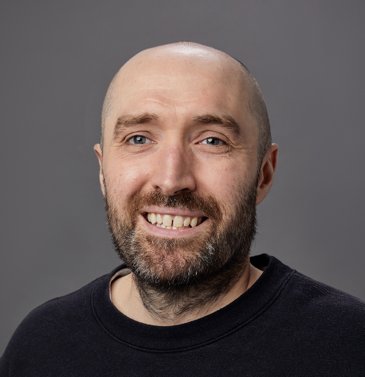 Close-up portrait of a smiling bald man with a beard, wearing a black t-shirt, against a gray background.