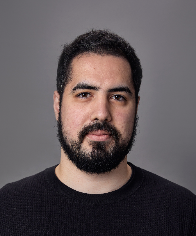 A portrait of a man with dark hair and a beard, wearing a black sweater, against a plain gray background.