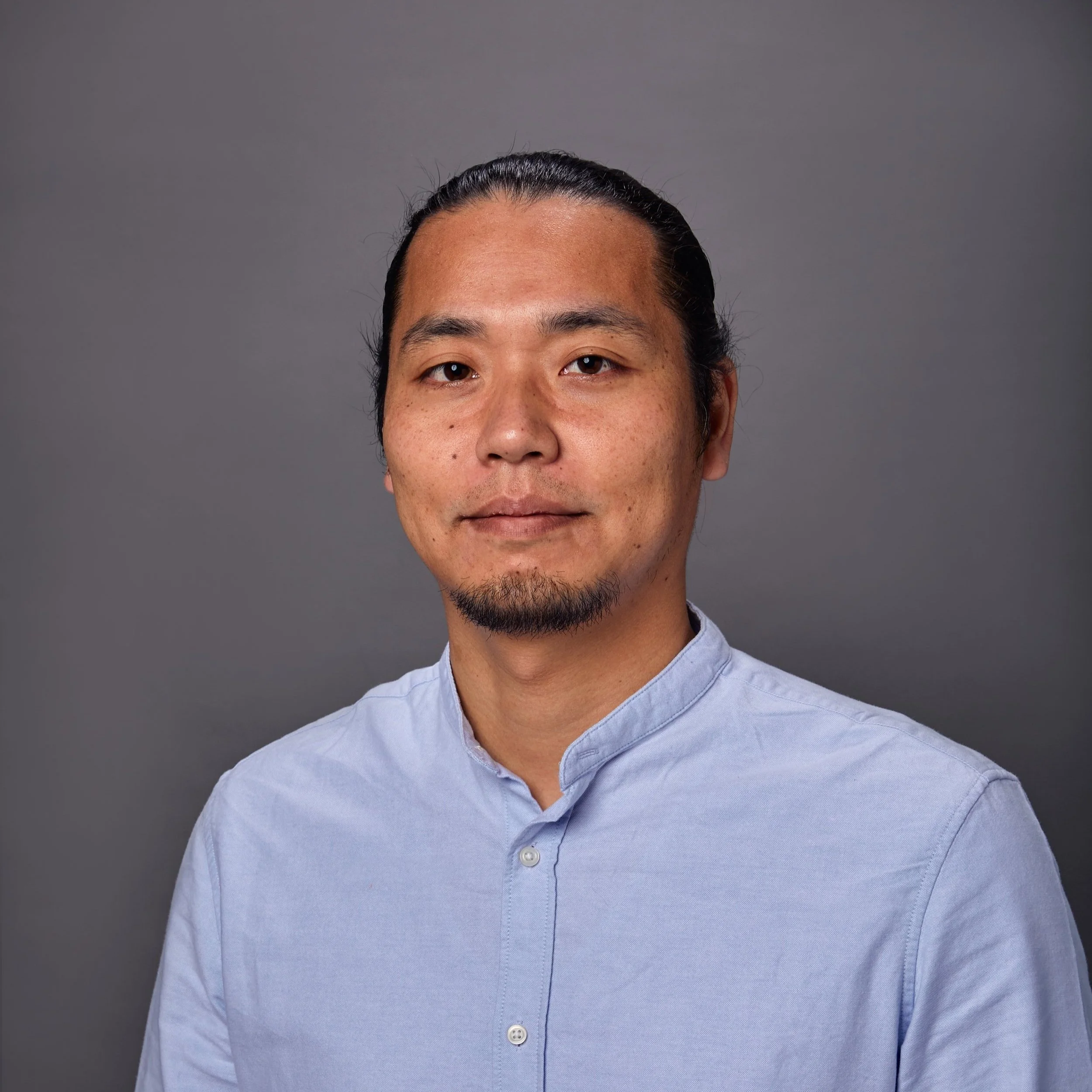 A man with dark hair tied back, a goatee, and mustache, wearing a light blue button-up shirt, against a gray background.