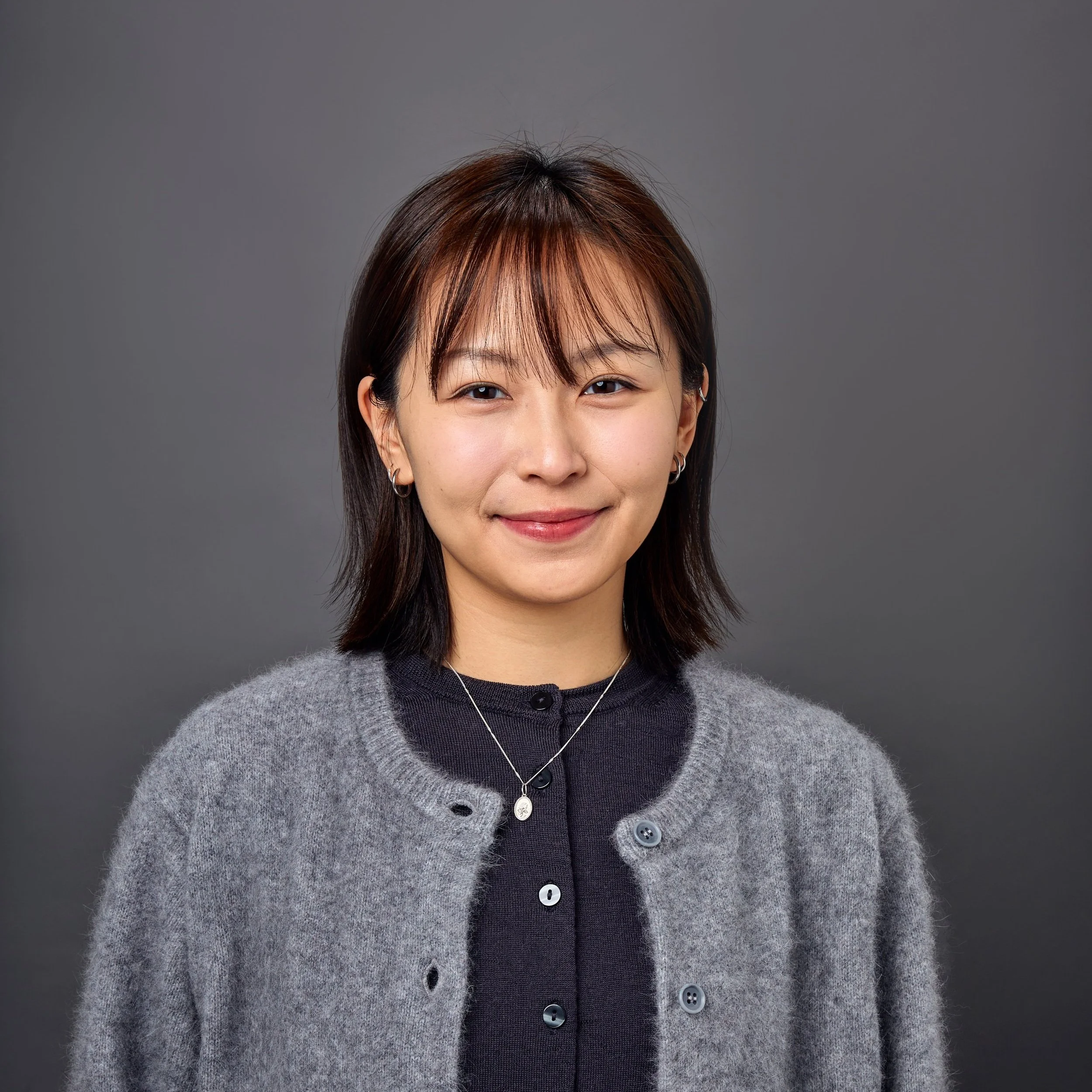 A young woman with shoulder-length dark brown hair, wearing a gray cardigan over a black top, standing against a gray background.