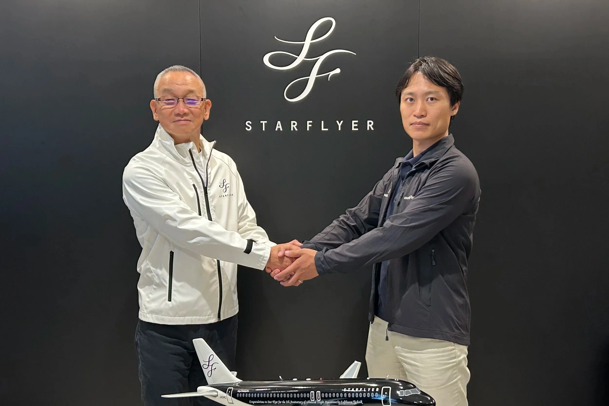 Two men shaking hands in front of a black wall with the logo 'Starflyer' and a model airplane on a table.