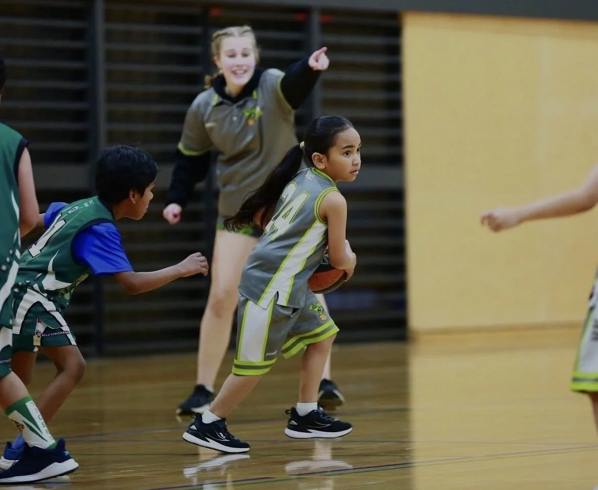 Young girls playing basketball in an indoor gym, one girl holding a basketball and looking to her right, others chasing or defending.