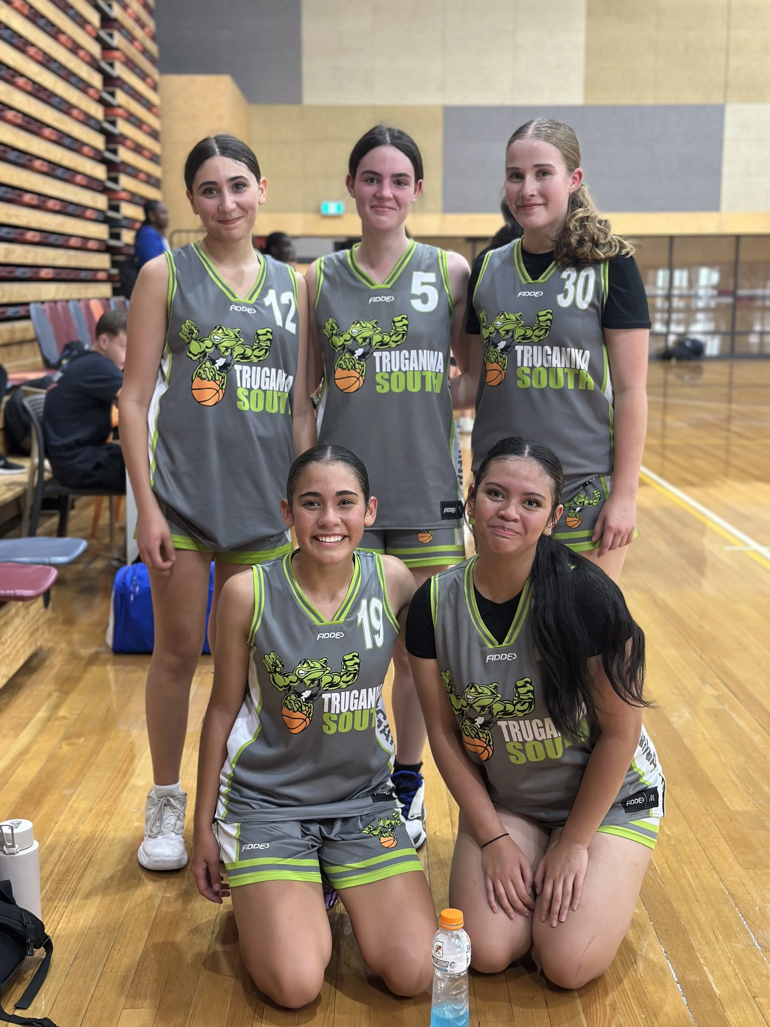 Six young female basketball players in gray and green uniforms with a frog mascot, posing on an indoor basketball court.