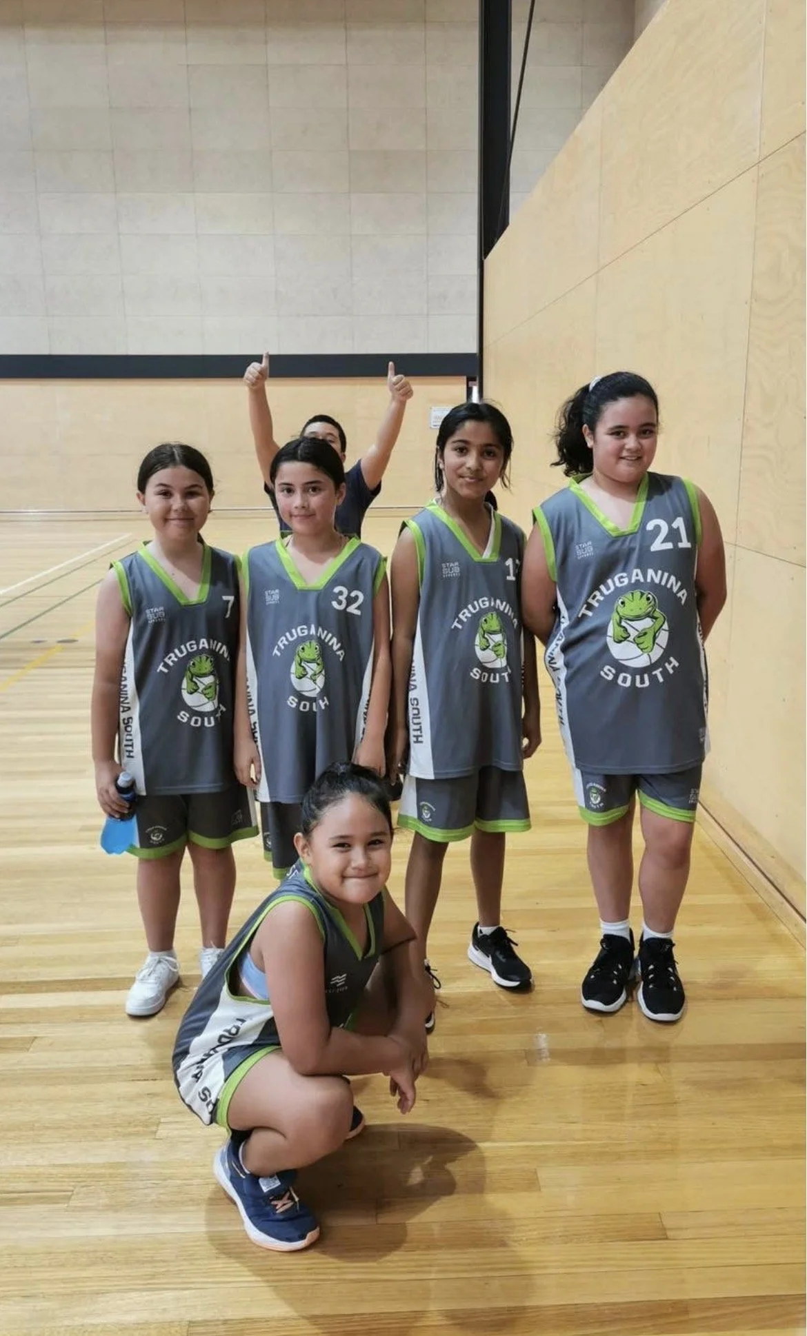 Group of six young girls in basketball uniforms in a gymnasium, some standing and one kneeling, smiling at the camera.