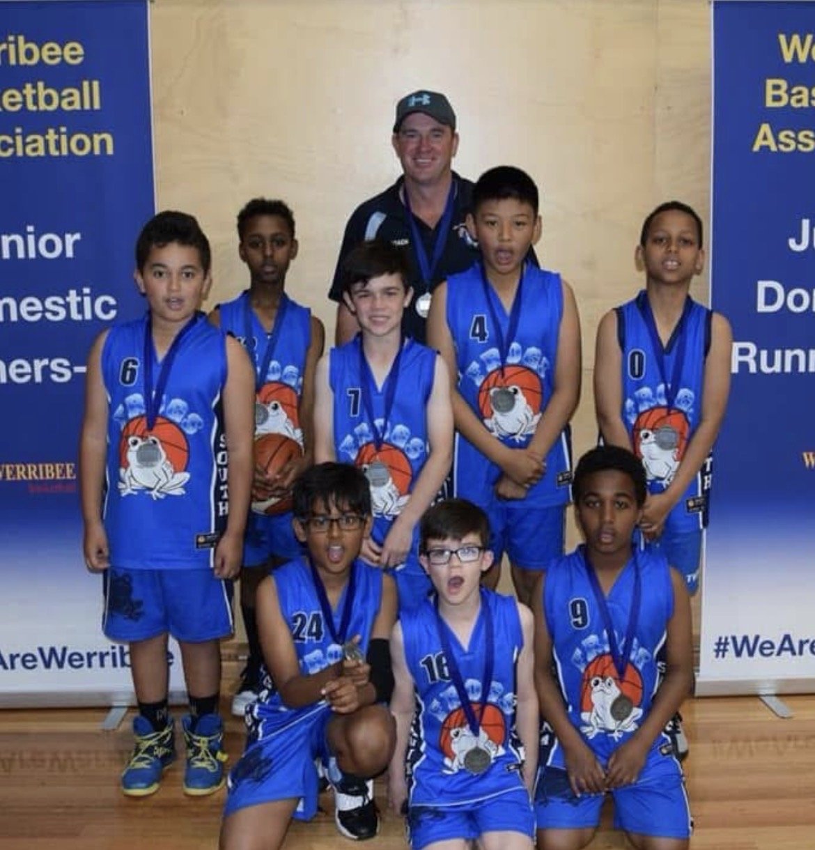 A youth basketball team in blue jerseys with a dog logo, wearing medals, posing with their coach after a game or tournament at a sports venue.