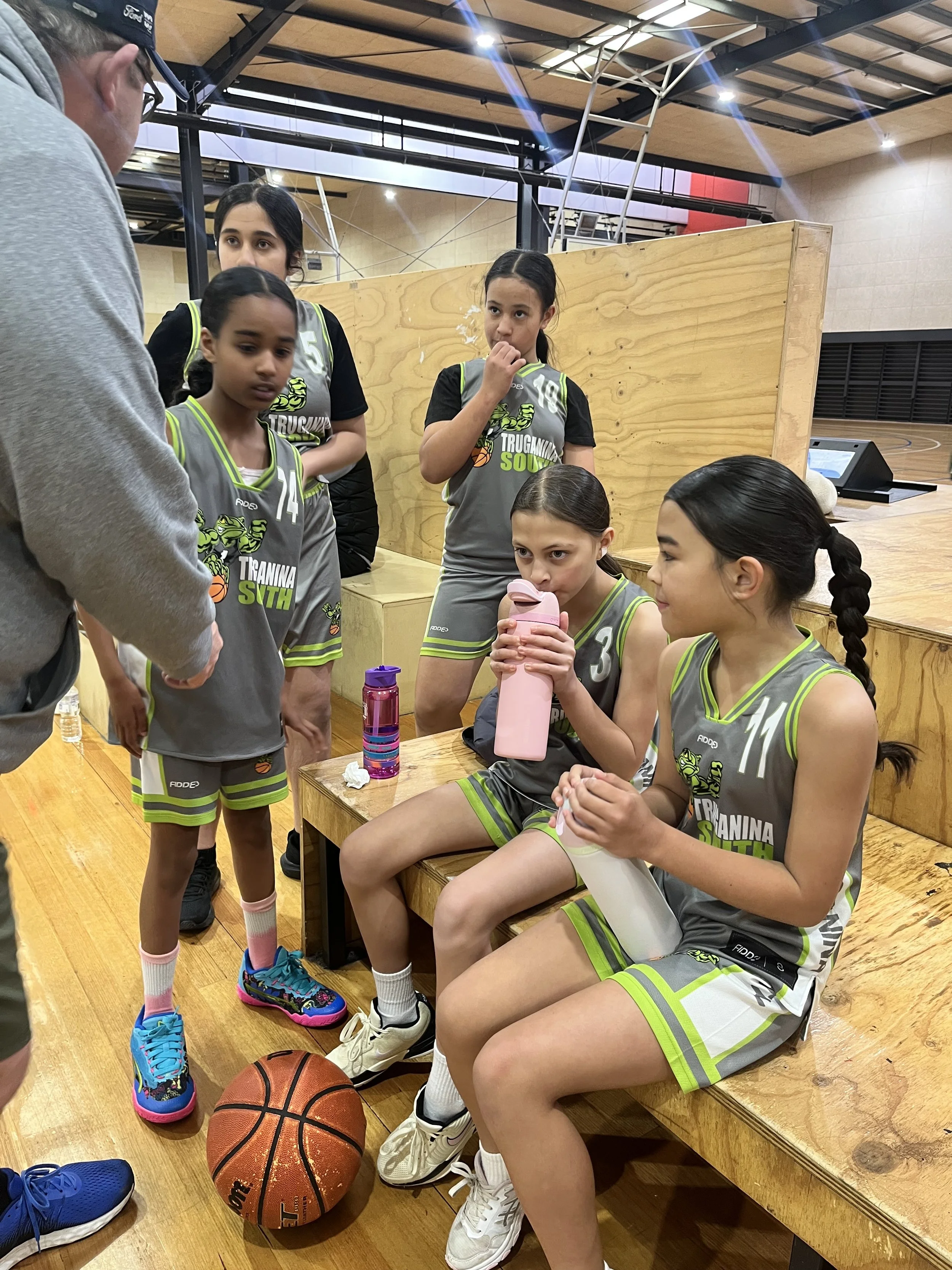 A group of young female basketball players in gray uniforms with green and white accents, sitting and standing on a wooden bench in a gymnasium, listening to their coach. One girl is drinking from a pink water bottle, and a basketball is on the floor