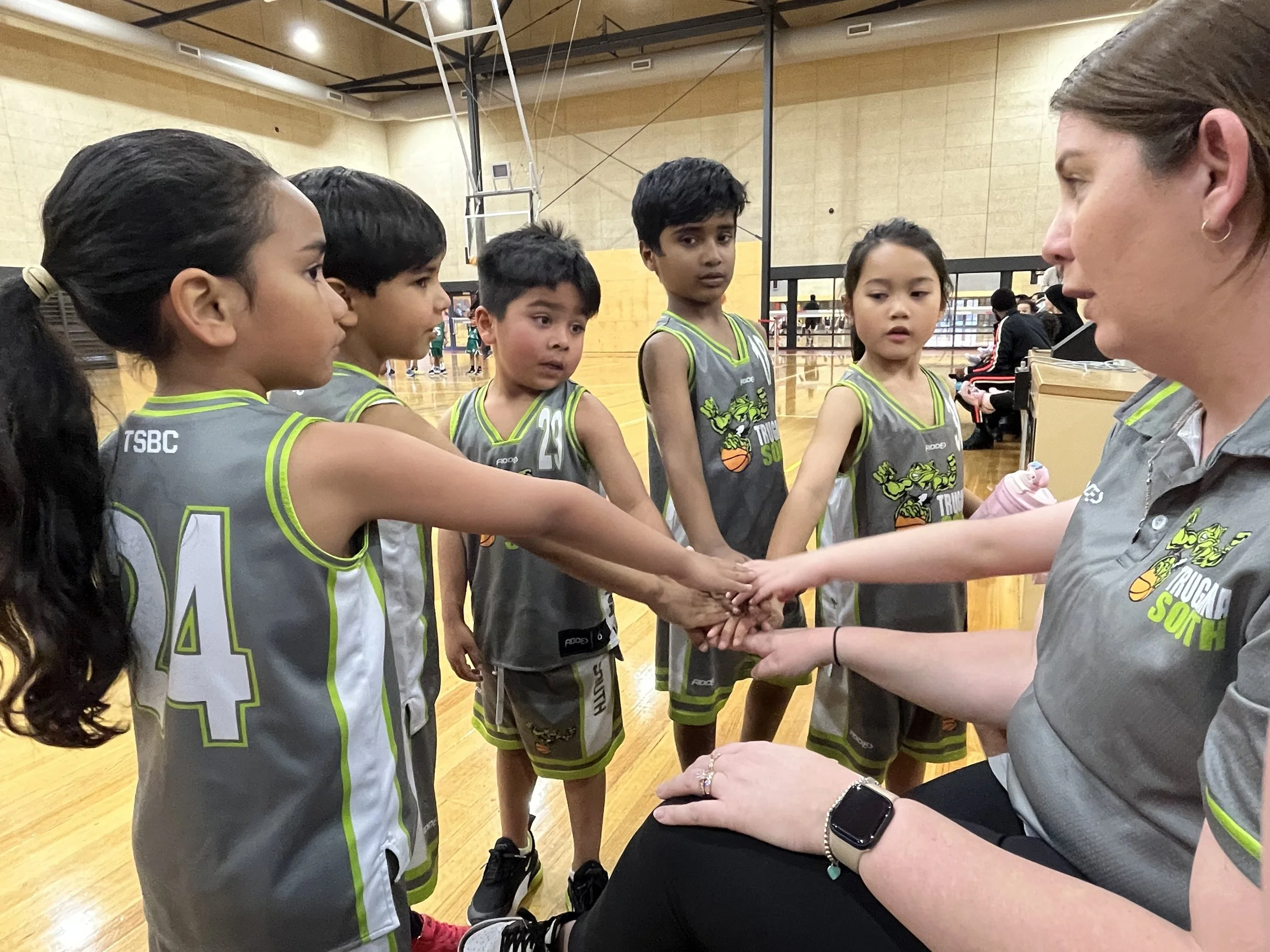 Young boys and girls in basketball uniforms huddle around a coach in a gym, placing their hands together for a team cheer.