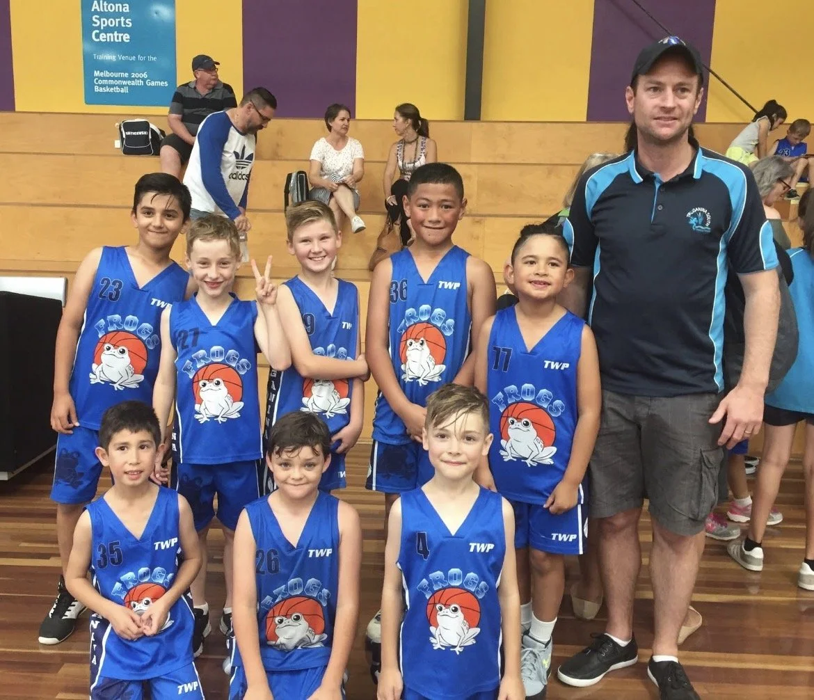 A group of young boys in blue basketball uniforms with a frog logo, standing with their coach in an indoor gymnasium. The boys are smiling and one is flashing a peace sign. There are spectators seated in the background.