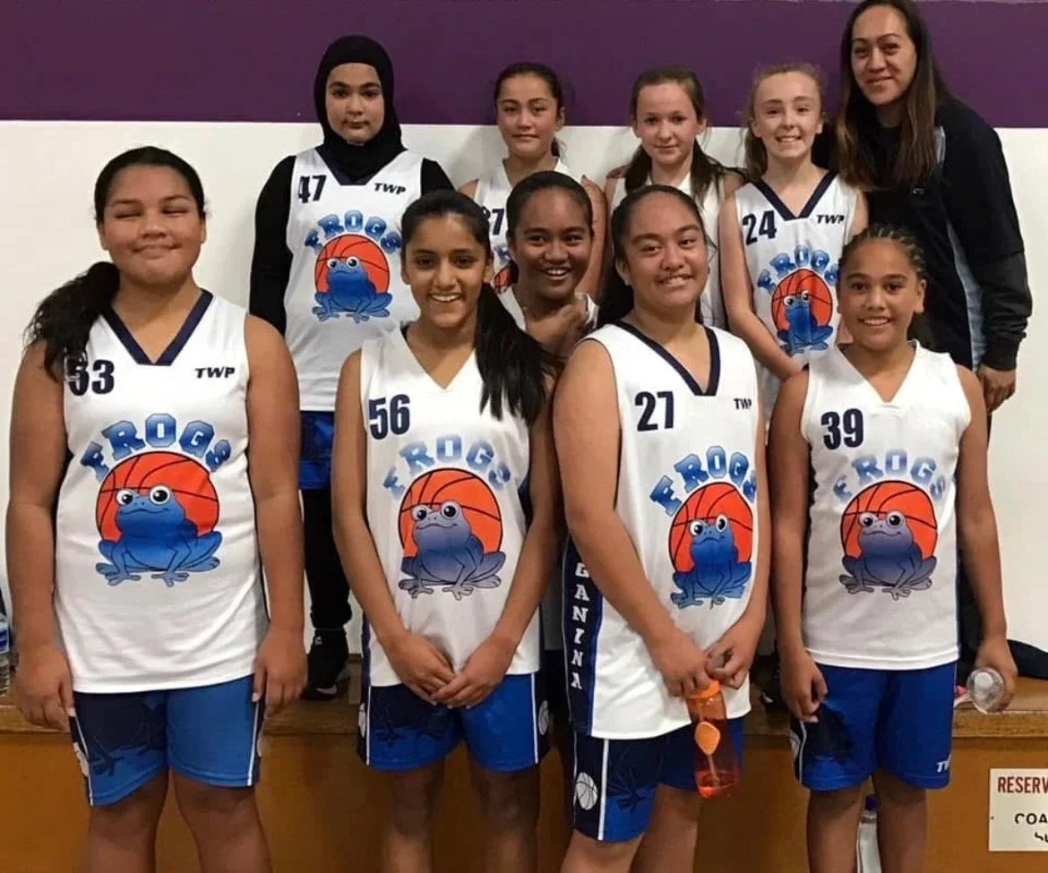 A girls' basketball team in white jerseys with blue shorts, featuring a frog logo, standing together with their coach in front of a purple and white wall.