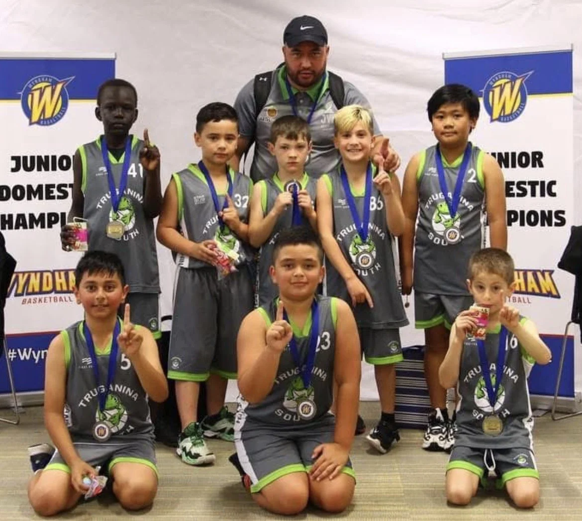 A youth basketball team with their coach, wearing gray and green uniforms and medals, posing with a trophy and flashing number one signs in front of a backdrop that reads 'Junior Domestic Champions'.